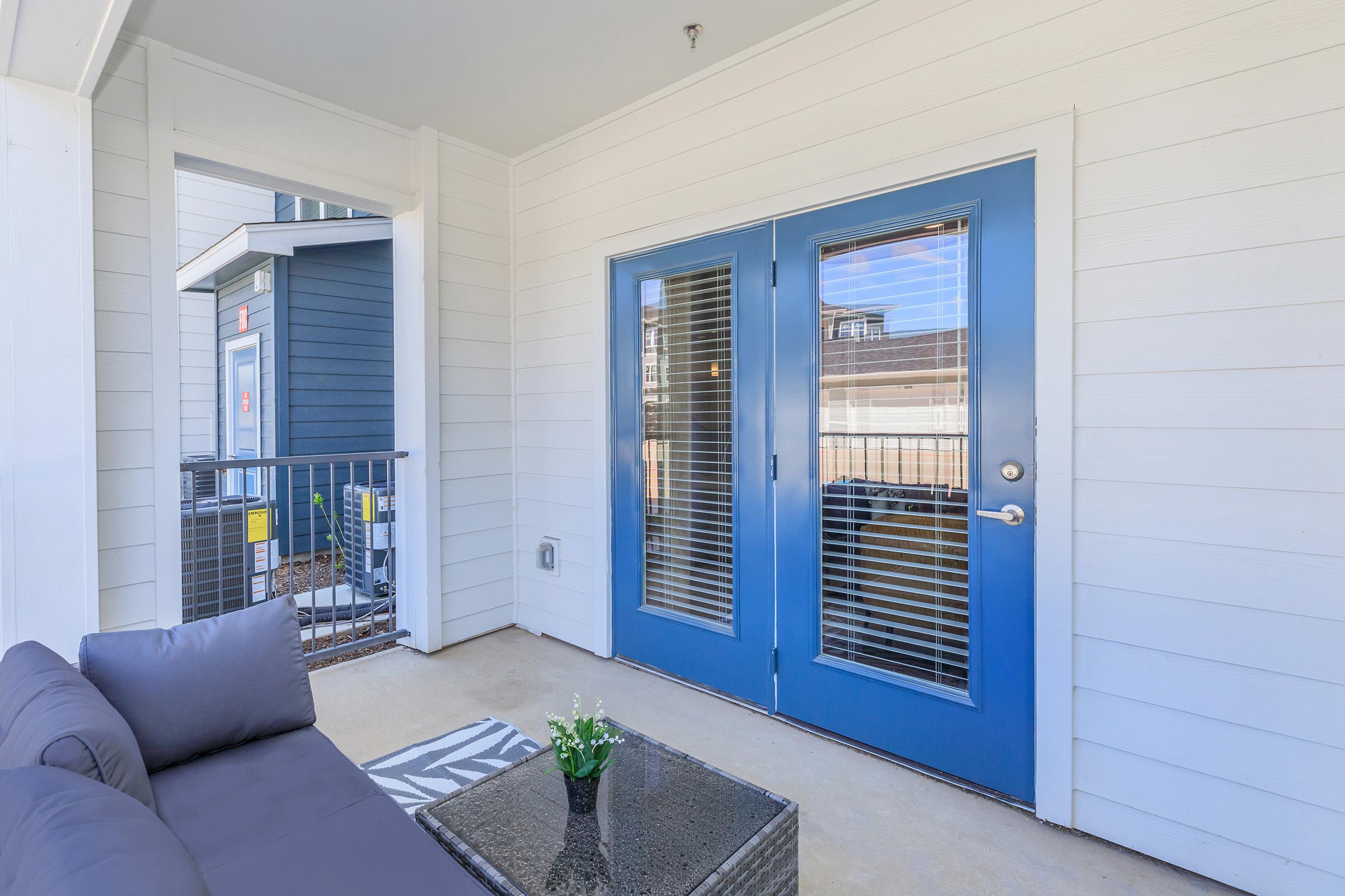A well-lit balcony area featuring a gray sofa and a small coffee table with a plant. Two blue double doors with blinds lead into an interior space, and there is a railing visible in the background. The walls are white, and the floor is concrete.