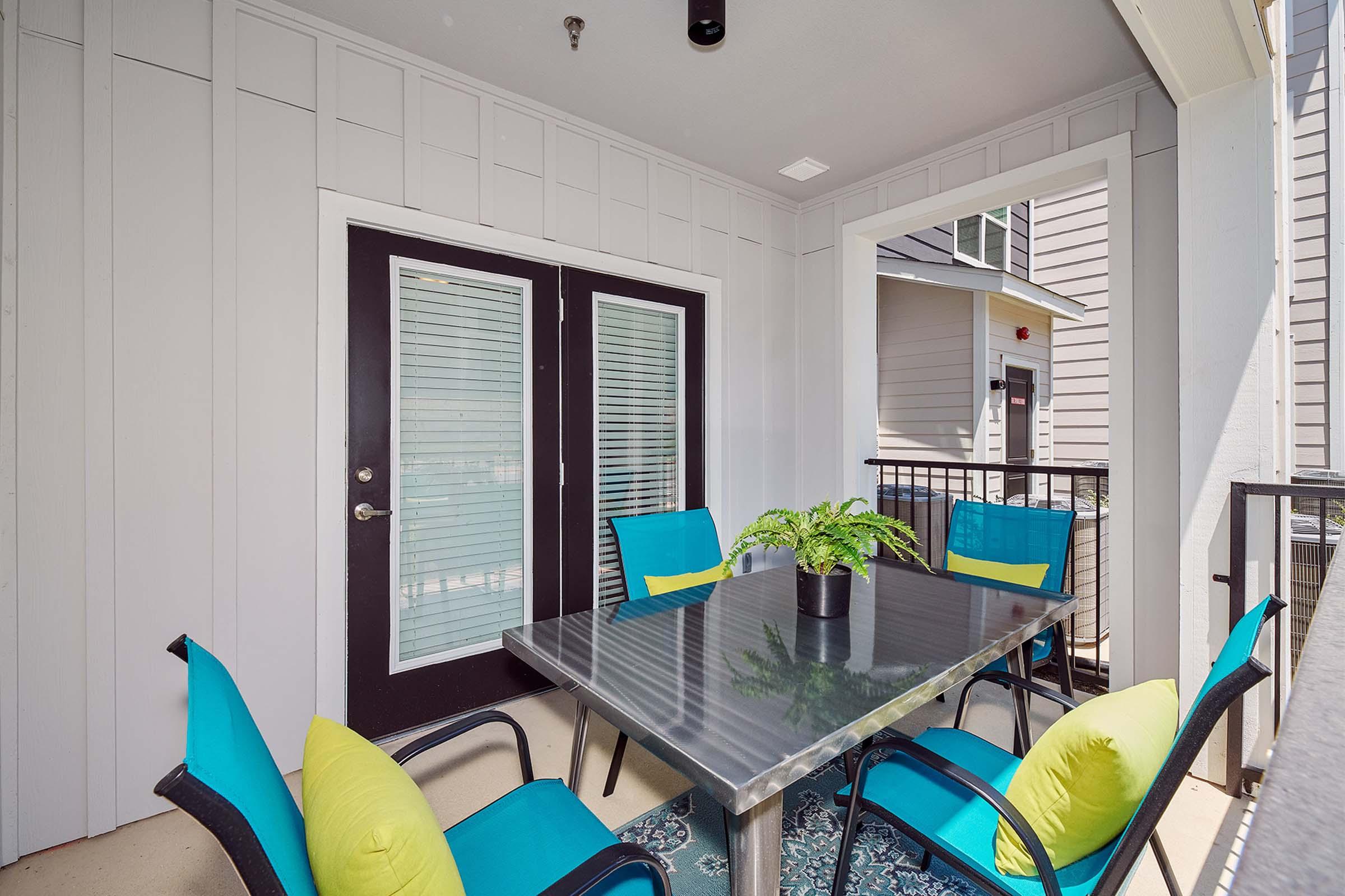 A modern outdoor patio with a table surrounded by six vibrant blue and green chairs. A small potted plant sits on the table. The background features a view of the building’s exterior with large doors and light-colored siding. The space is well-lit and inviting, perfect for outdoor gatherings.