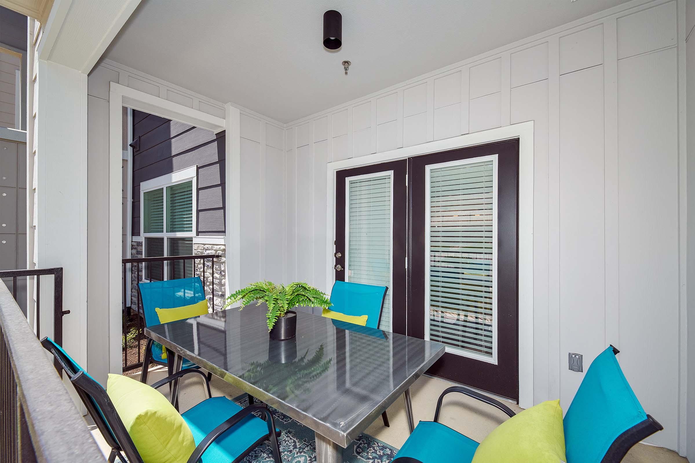 A cozy outdoor patio area featuring a glass table surrounded by four bright blue chairs with green cushions. The backdrop includes a gray wooden wall and several windows, creating a welcoming and modern ambiance. A potted plant sits in the center of the table, enhancing the inviting atmosphere.