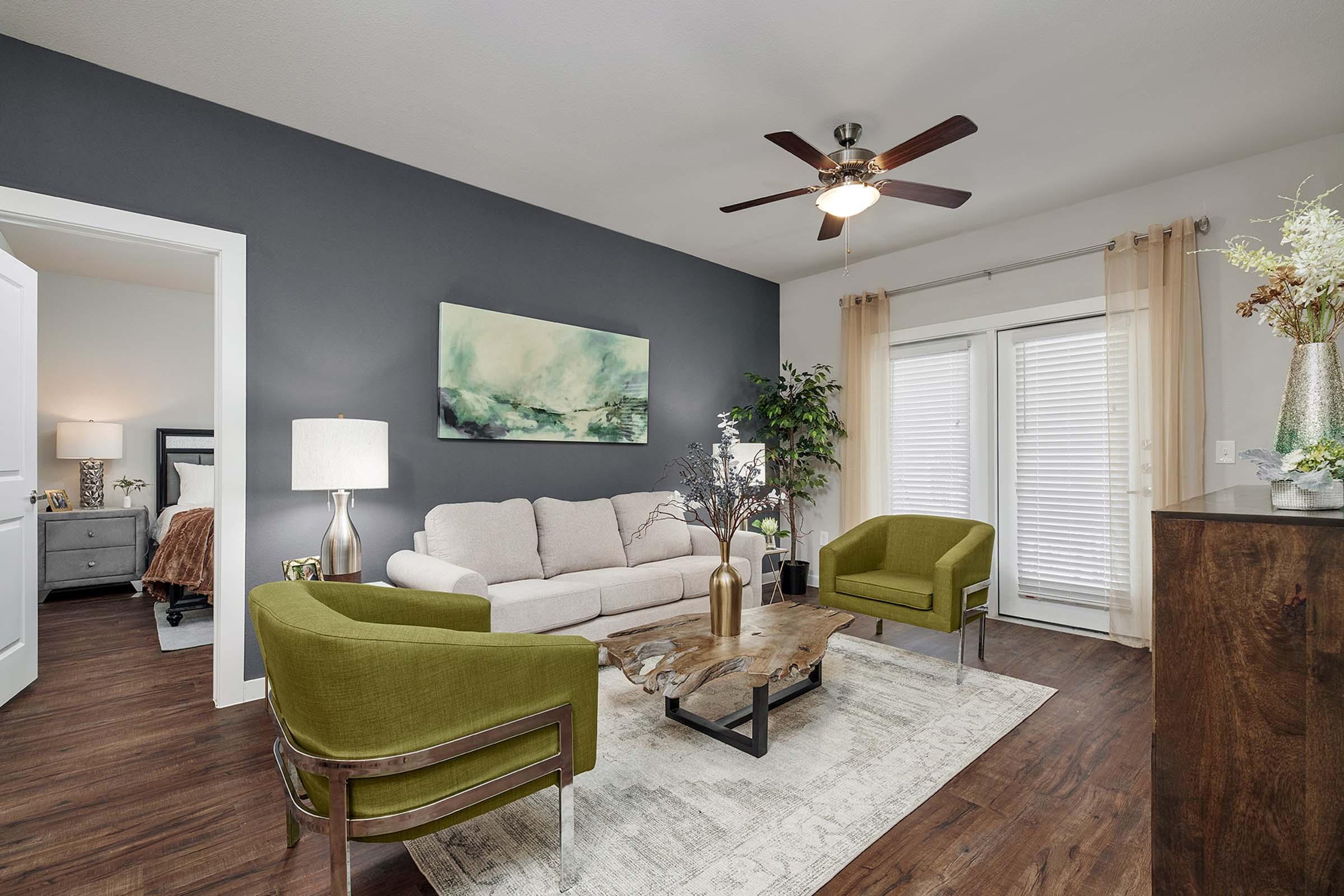 A modern living room featuring a light beige sofa, two green armchairs, and a wooden coffee table. There's a decorative plant in the corner, a floor lamp, and wall art. The room has large windows with sheer curtains, allowing natural light to illuminate the space. Hardwood flooring adds warmth to the design.