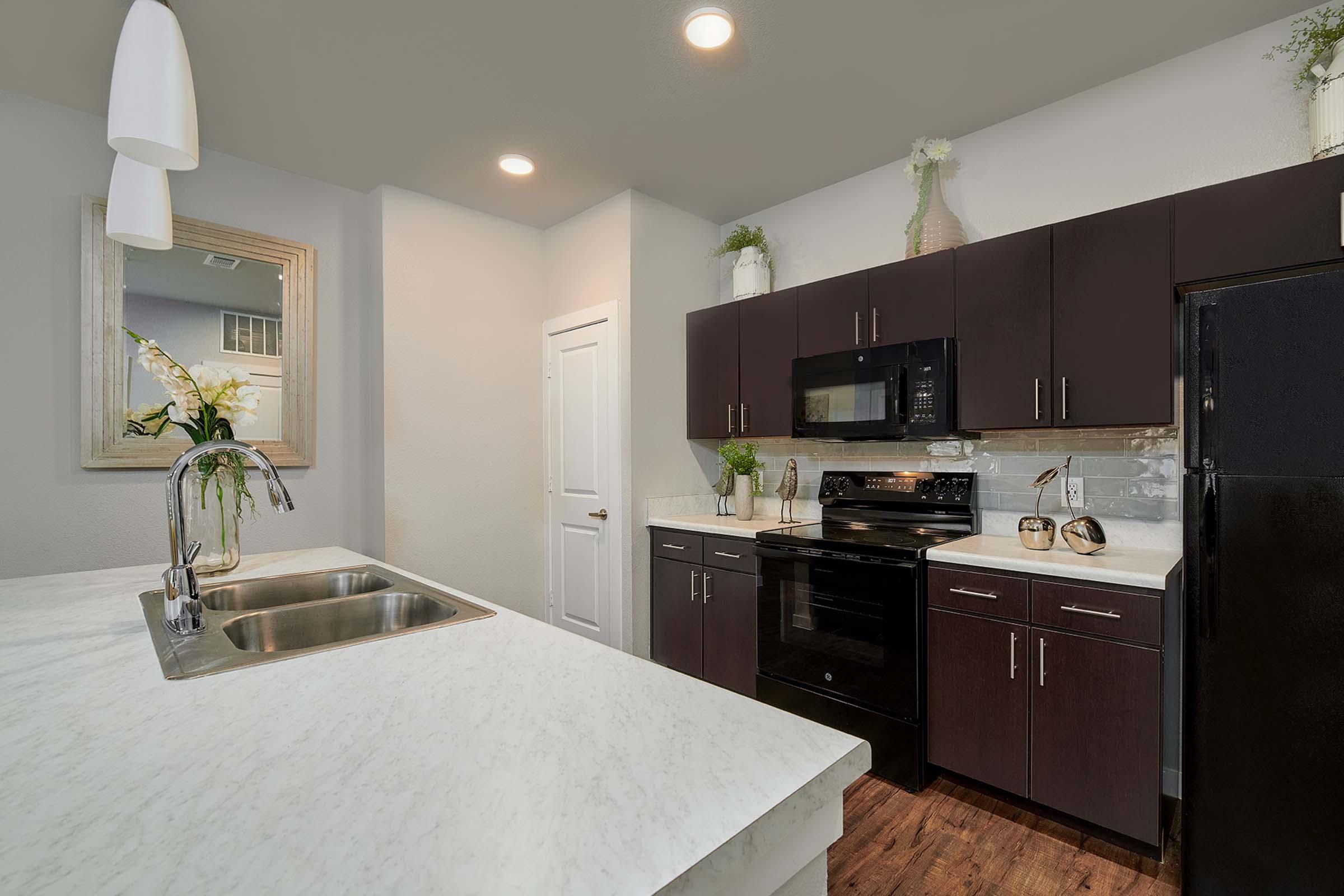 Modern kitchen featuring dark cabinetry, a black stove, microwave, and refrigerator. A white marble countertop with a double sink is positioned in the foreground. The space is well-lit with recessed ceiling lights, and decorative plants enhance the aesthetic. A large mirror reflects light, adding depth to the room.