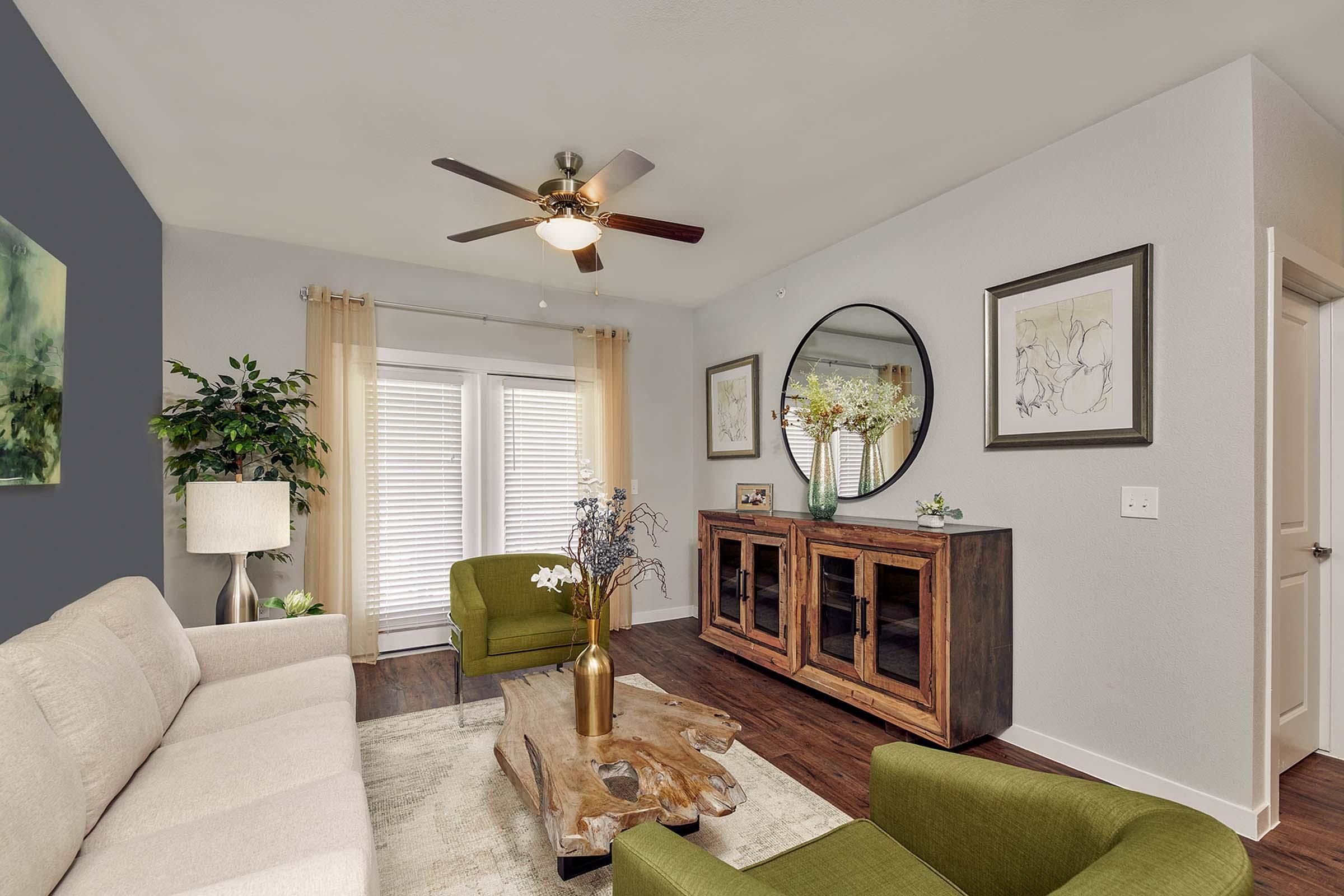 A modern living room featuring a light-colored sofa, two green chairs, and a wooden coffee table. There is a round mirror on the wall, decorative art, and a stylish cabinet against the wall. Sunlight filters through the windows, and potted plants add a touch of greenery. The walls are painted a soft gray.