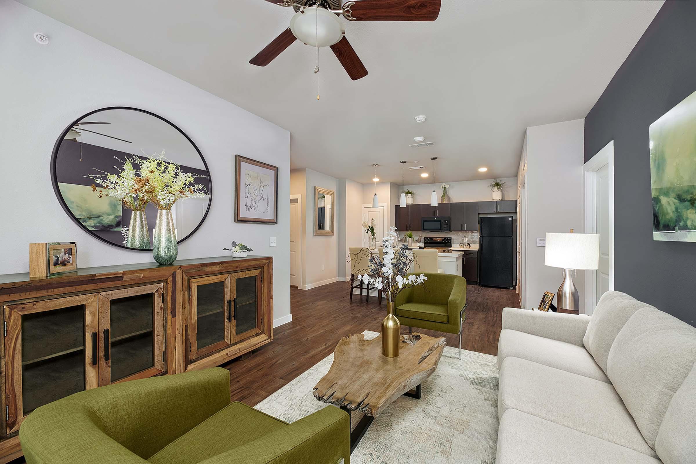 A modern living room showcasing a cozy arrangement with a light-colored sofa and green chairs. There is a wooden coffee table in the center, surrounded by decorative items. The room features a large circular mirror, wall art, and a TV unit against a dark accent wall, with warm lighting and a ceiling fan above.