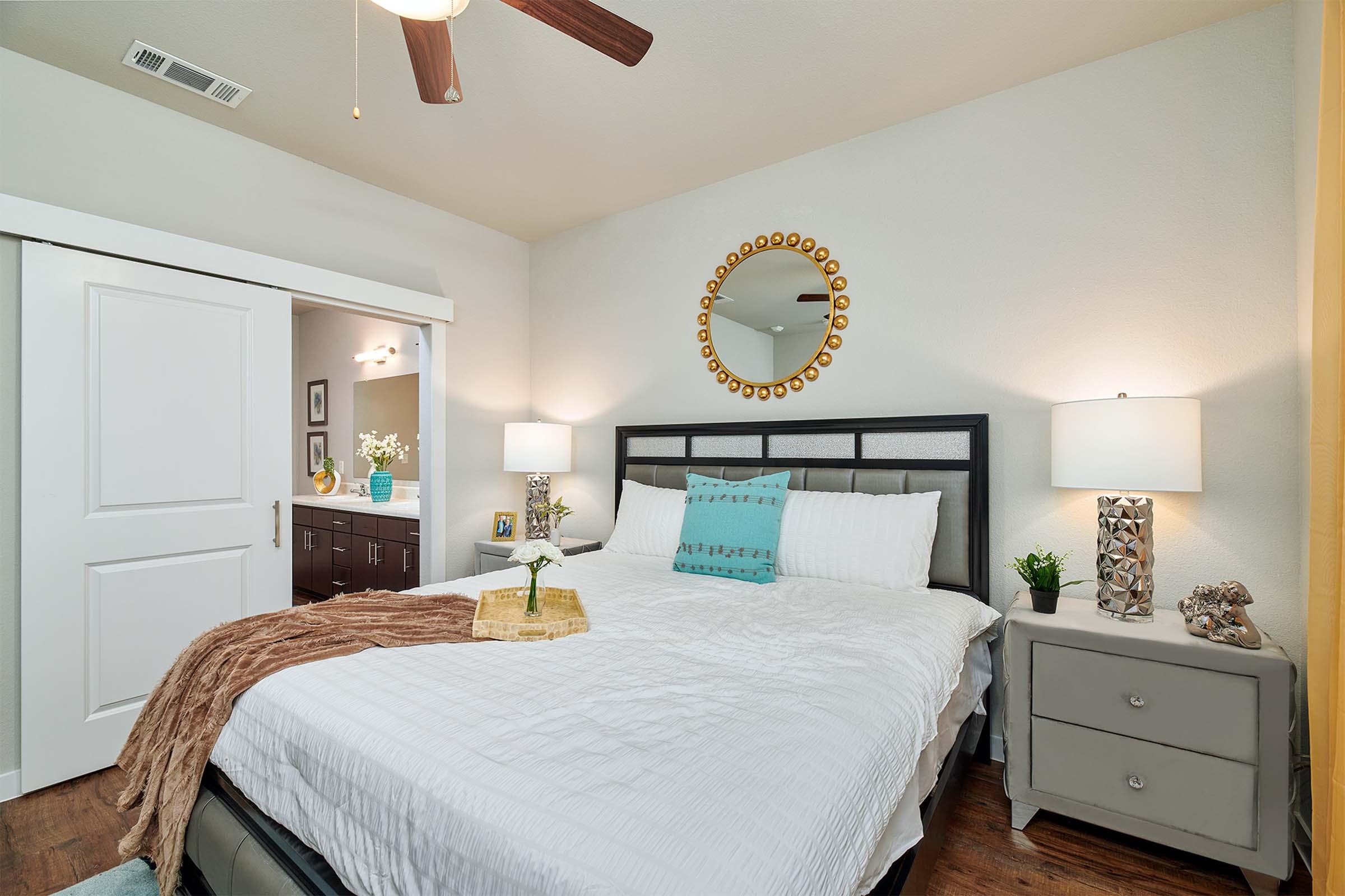 A cozy bedroom featuring a gray bed with white bedding and decorative pillows. There is a wooden dresser and two stylish lamps on bedside tables. A large round mirror is hanging above the bed, and a textured throw blanket is draped over the bed. A doorway leads to an adjoining bathroom area.