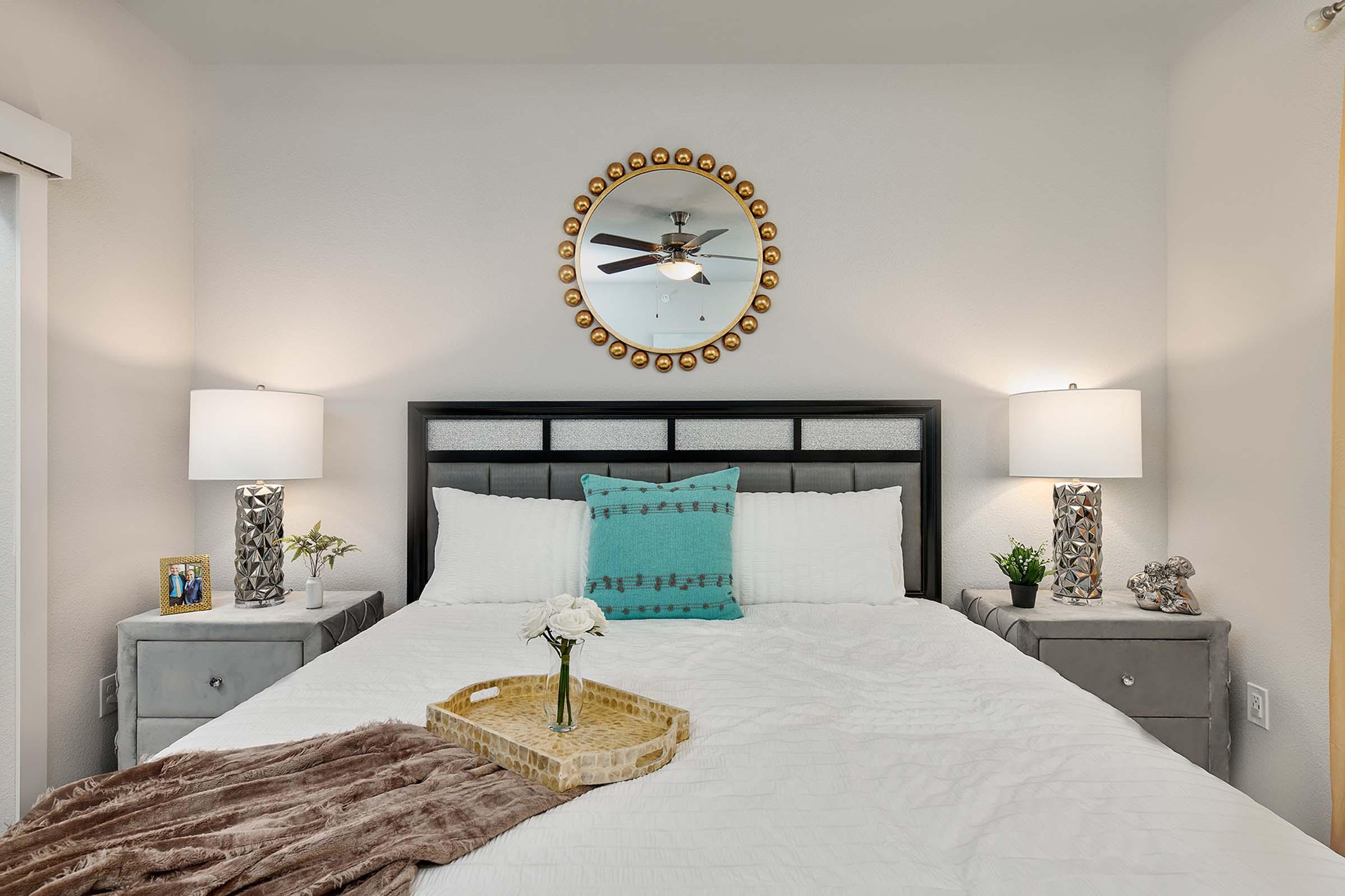 A cozy bedroom featuring a neatly made bed with white linens and a turquoise decorative pillow. On either side of the bed, there are modern nightstands with stylish lamps. A round mirror with a gold border hangs above the headboard, and a decorative tray with flowers sits on the bed, adding a touch of elegance.