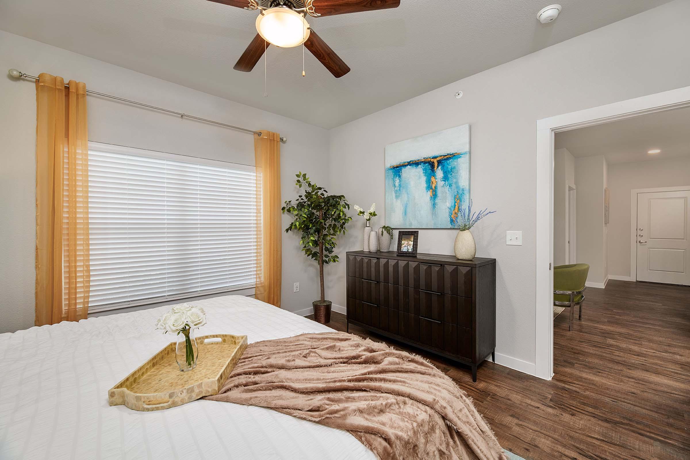 Bright, cozy bedroom featuring a light-colored bed with a decorative throw, a wooden dresser adorned with a vase and framed art, large window with sheer curtains, and a potted plant. Warm natural light enhances the inviting atmosphere, with a door leading to another room visible in the background.