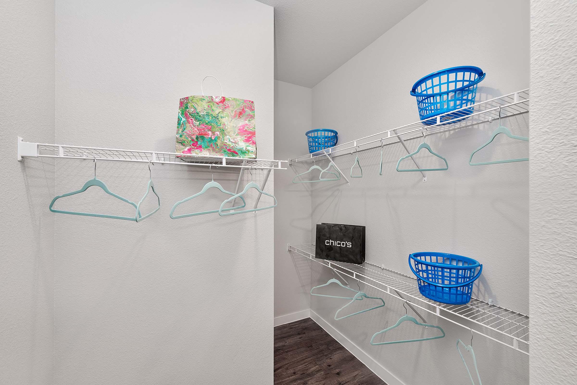 A neatly organized closet featuring white wire shelves with several light blue hangers, two blue laundry baskets, and a decorative bag. The walls are painted light gray, and the flooring is dark wood, creating a clean and spacious look.