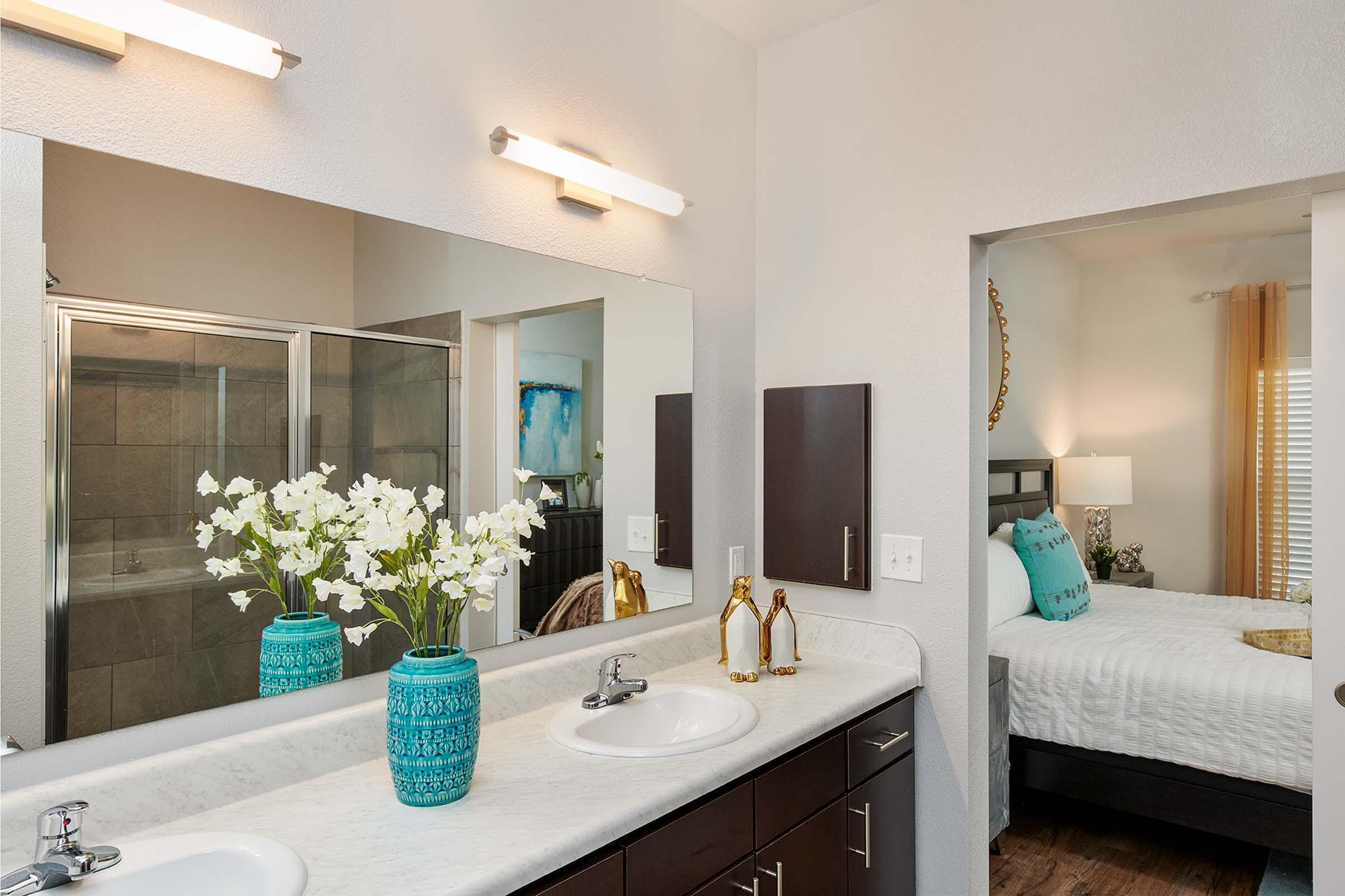 A modern bathroom featuring double sinks with a marble countertop, a decorative blue vase filled with white flowers, and stylish faucets. A large mirror reflects the space, including an adjacent room with a bed and decorative pillows, enhancing the overall contemporary aesthetic.