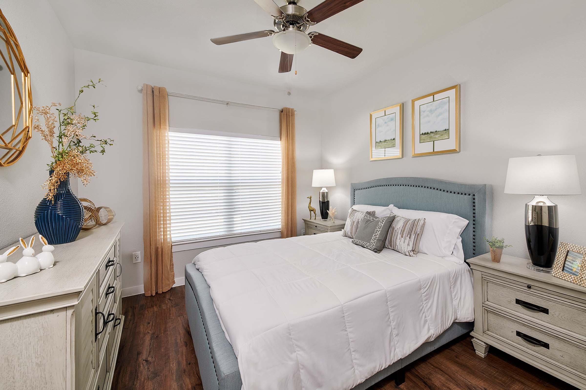 A cozy bedroom featuring a queen-sized bed with white bedding and decorative pillows. The room includes a stylish blue upholstered headboard, two lamps on nightstands, a large window with sheer curtains, and framed artwork on the wall. A dresser with decorative accents is also visible, creating a warm and inviting atmosphere.