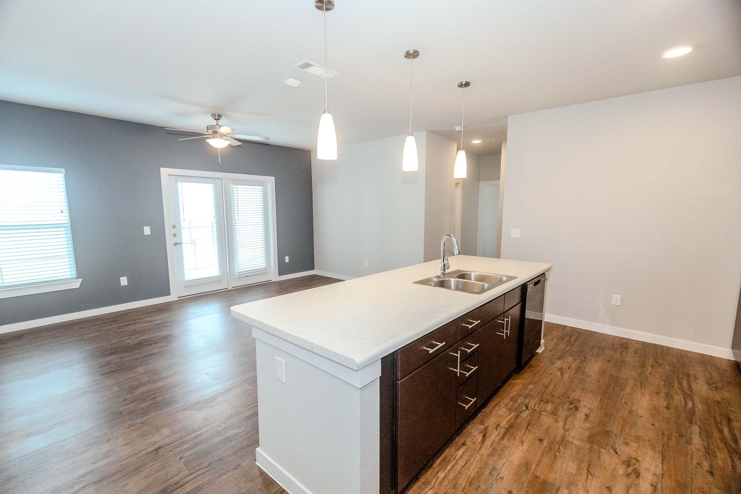 A modern, open-concept living space featuring a kitchen island with a sink, dark cabinetry, and pendant lighting. The room has light wood flooring and gray walls, with a ceiling fan and large windows letting in natural light. Two glass doors lead to an outdoor area.