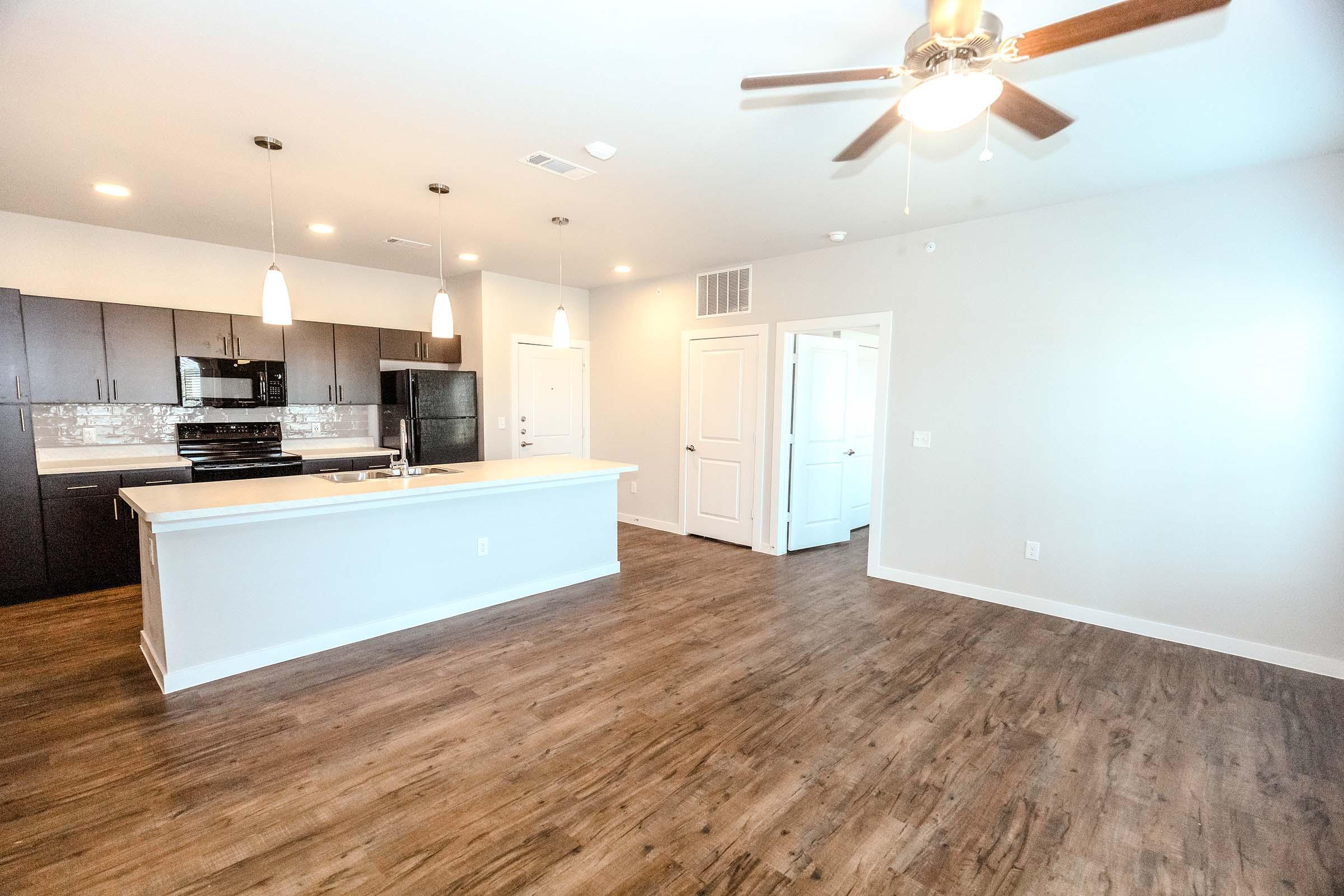 A modern interior of an open-concept living space featuring a kitchen with dark cabinetry, a white countertop, and stainless steel appliances. The room has a ceiling fan, pendant lighting, and hardwood flooring, with a doorway leading to another room. Natural light brightens the space.