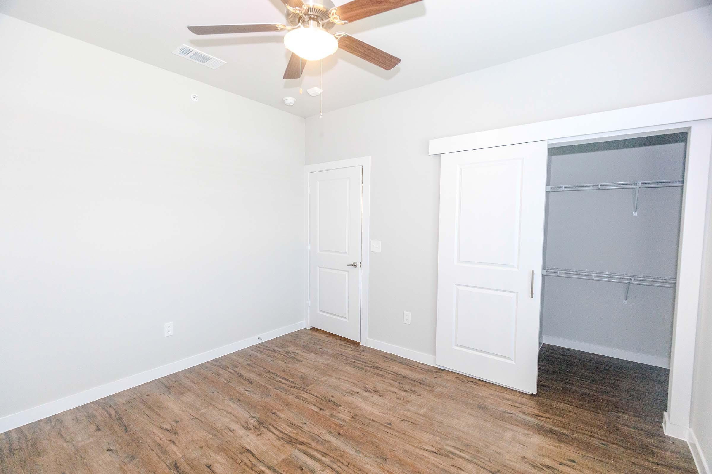 A spacious, empty room with light-colored walls and a ceiling fan. The floor features wood-like laminate, and there is a closet with a sliding door, revealing shelves inside. Natural light enters the room, creating a bright and airy atmosphere.