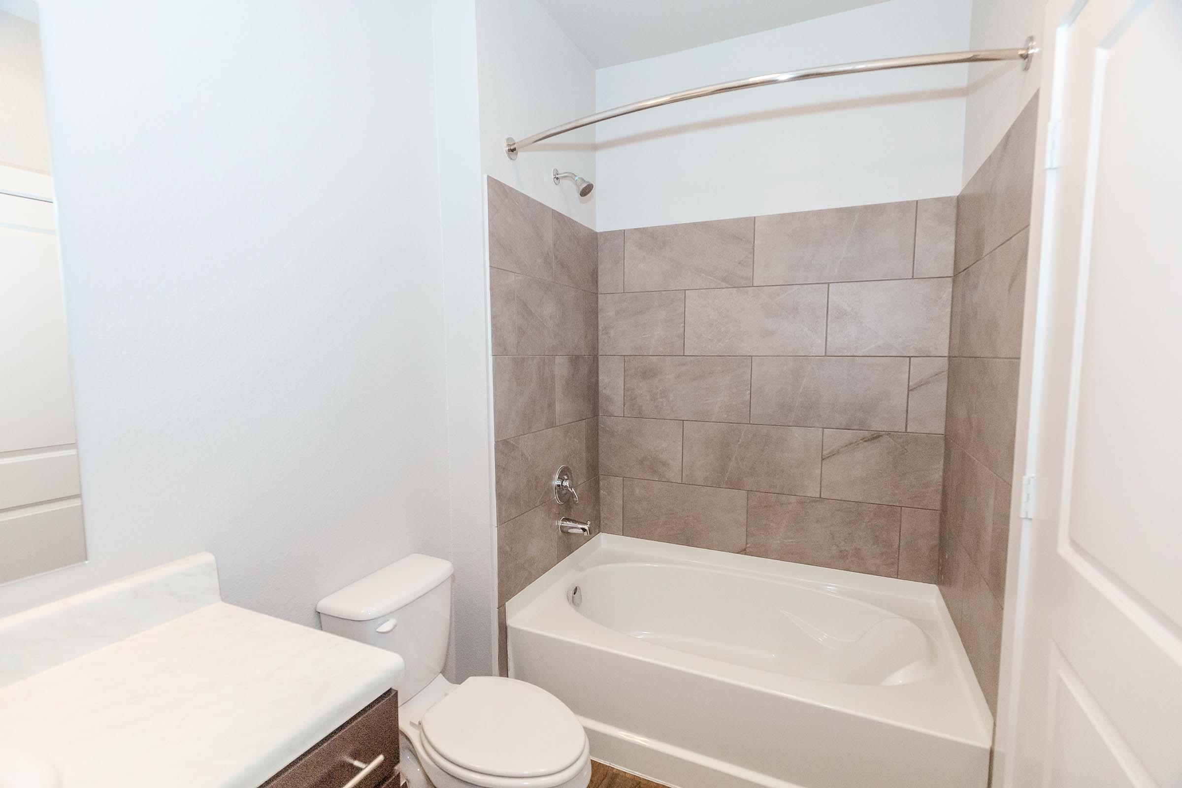 A clean, modern bathroom featuring a white bathtub with a shower curtain, tiled walls in neutral tones, a white toilet, and a marble countertop with a sink. The walls are painted light colors, providing a bright and airy feel.