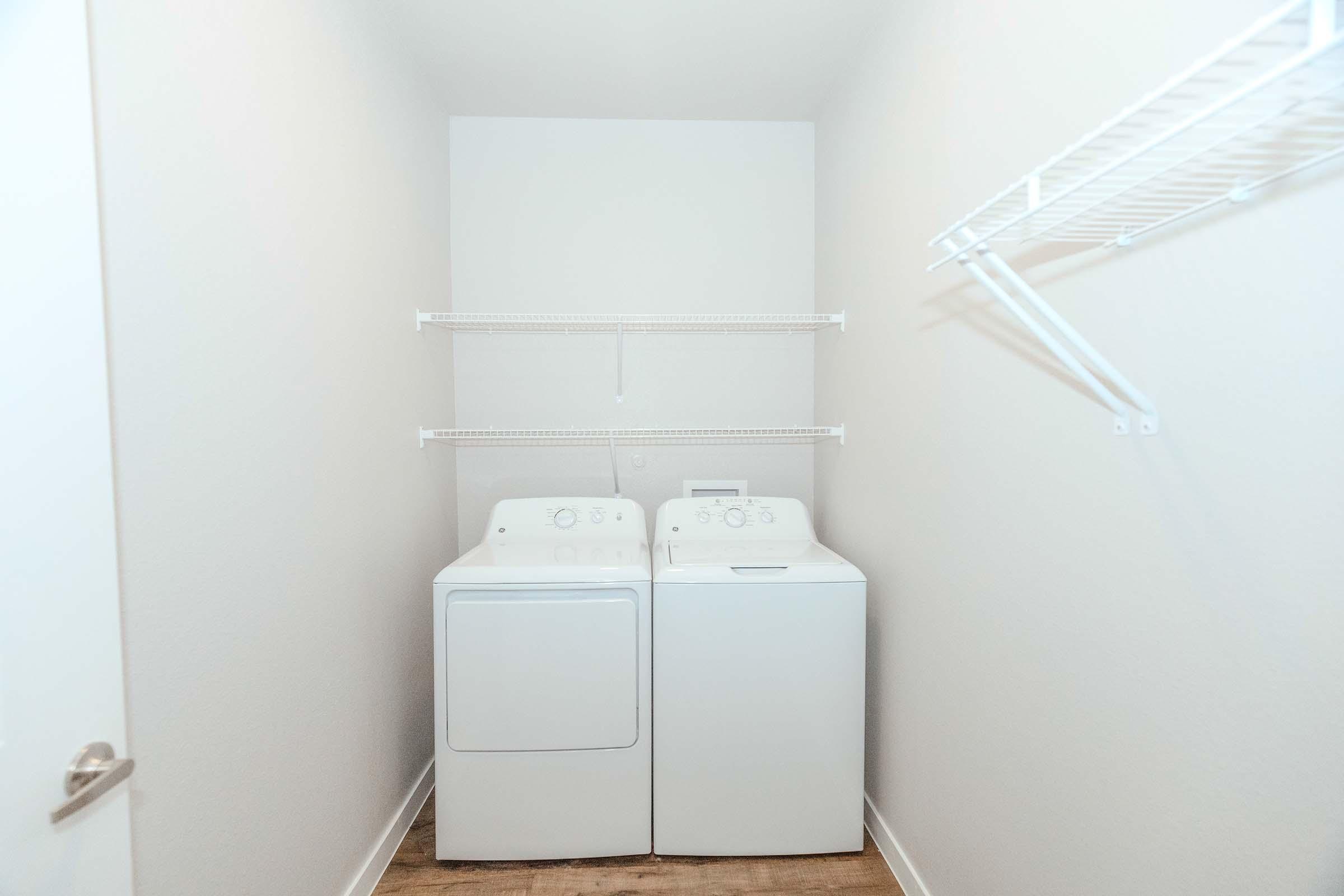 A clean laundry room featuring a white washing machine and dryer side by side against a light-colored wall. The room has wire shelving above the appliances and a single clothes rack on the right side, with light wood flooring adding a warm touch to the space.