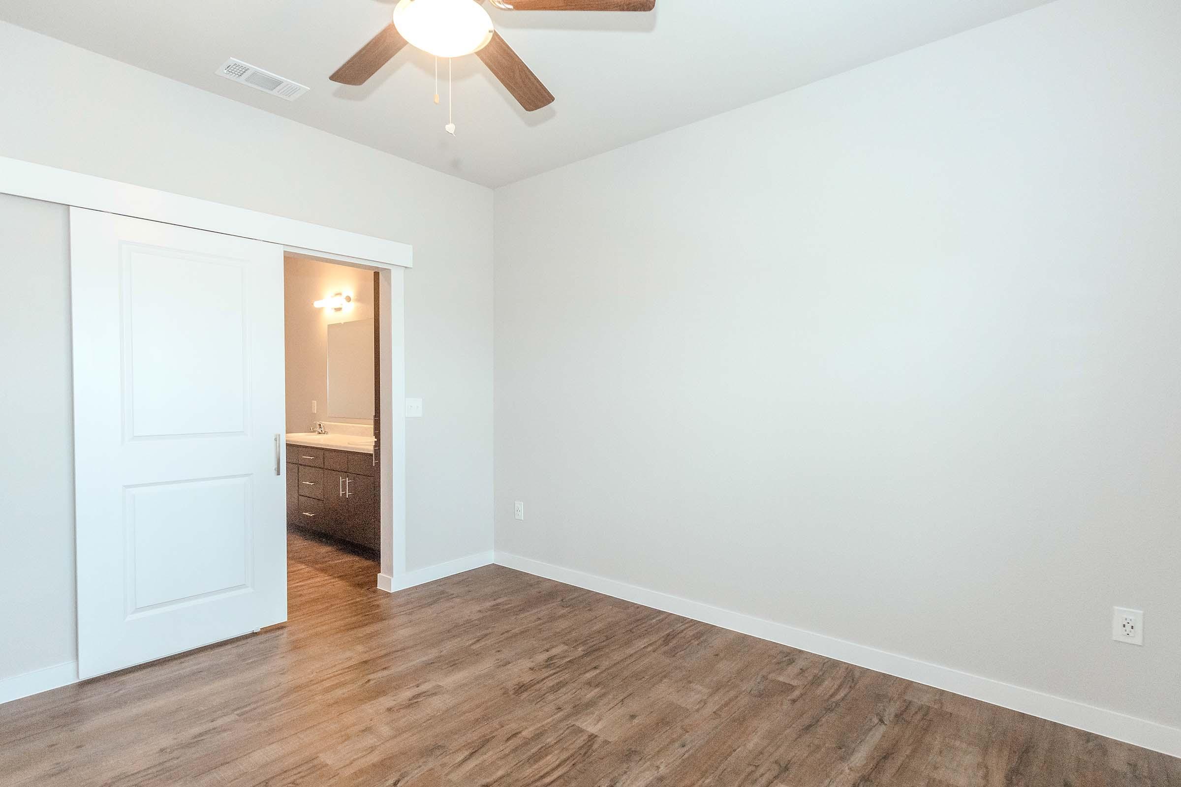 A bright, empty room with light-colored walls and wooden flooring. A sliding closet door is visible on the left, leading to a bathroom with modern fixtures. The ceiling features a ceiling fan, and there is ample natural light entering the space, creating a clean and airy atmosphere.