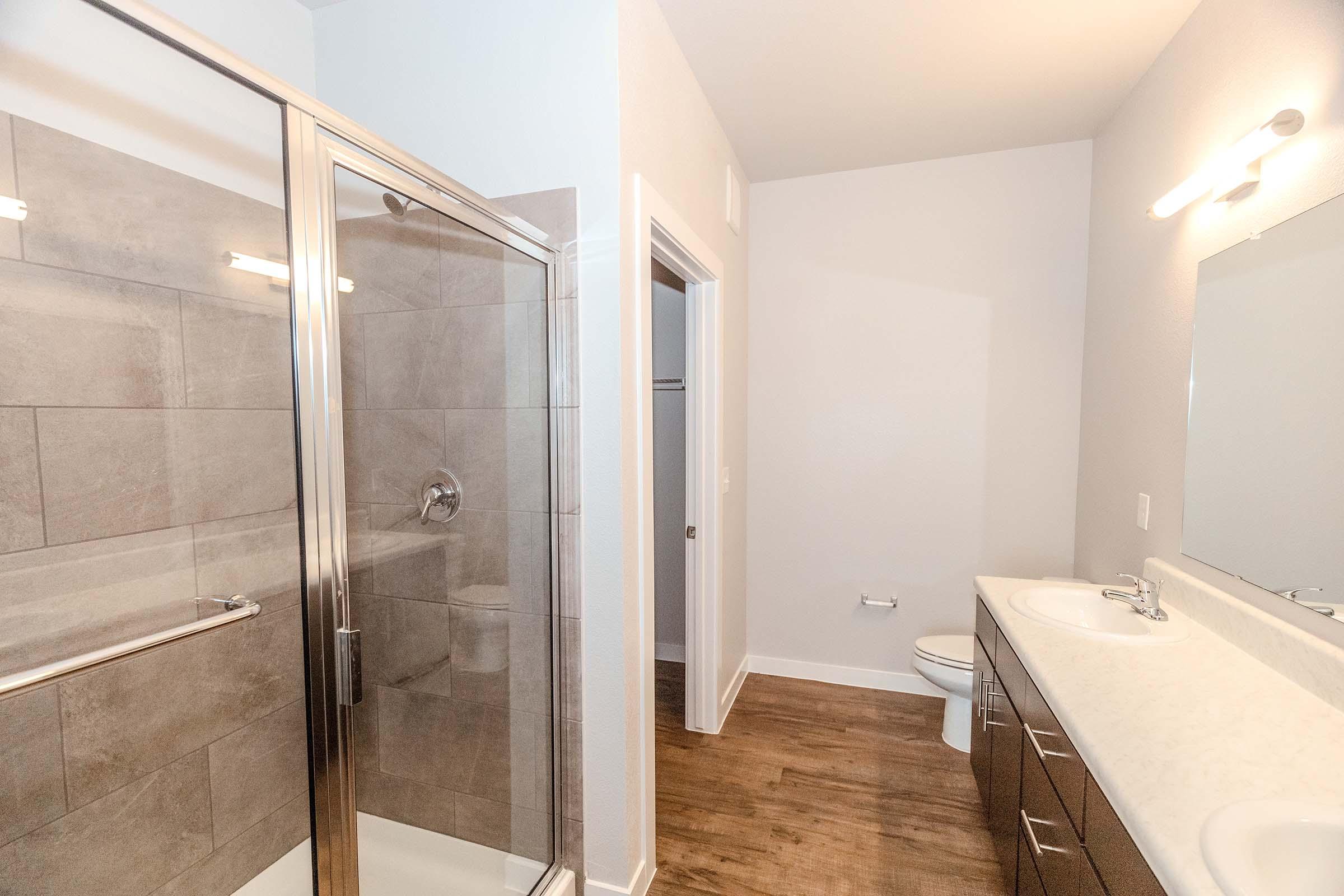 A modern bathroom featuring a glass-enclosed shower, double sink vanity with a countertop, mirrored wall, and a toilet. The walls are painted light gray, and the floor has a wood-like finish. Natural light brightens the space, which has a clean and contemporary design.