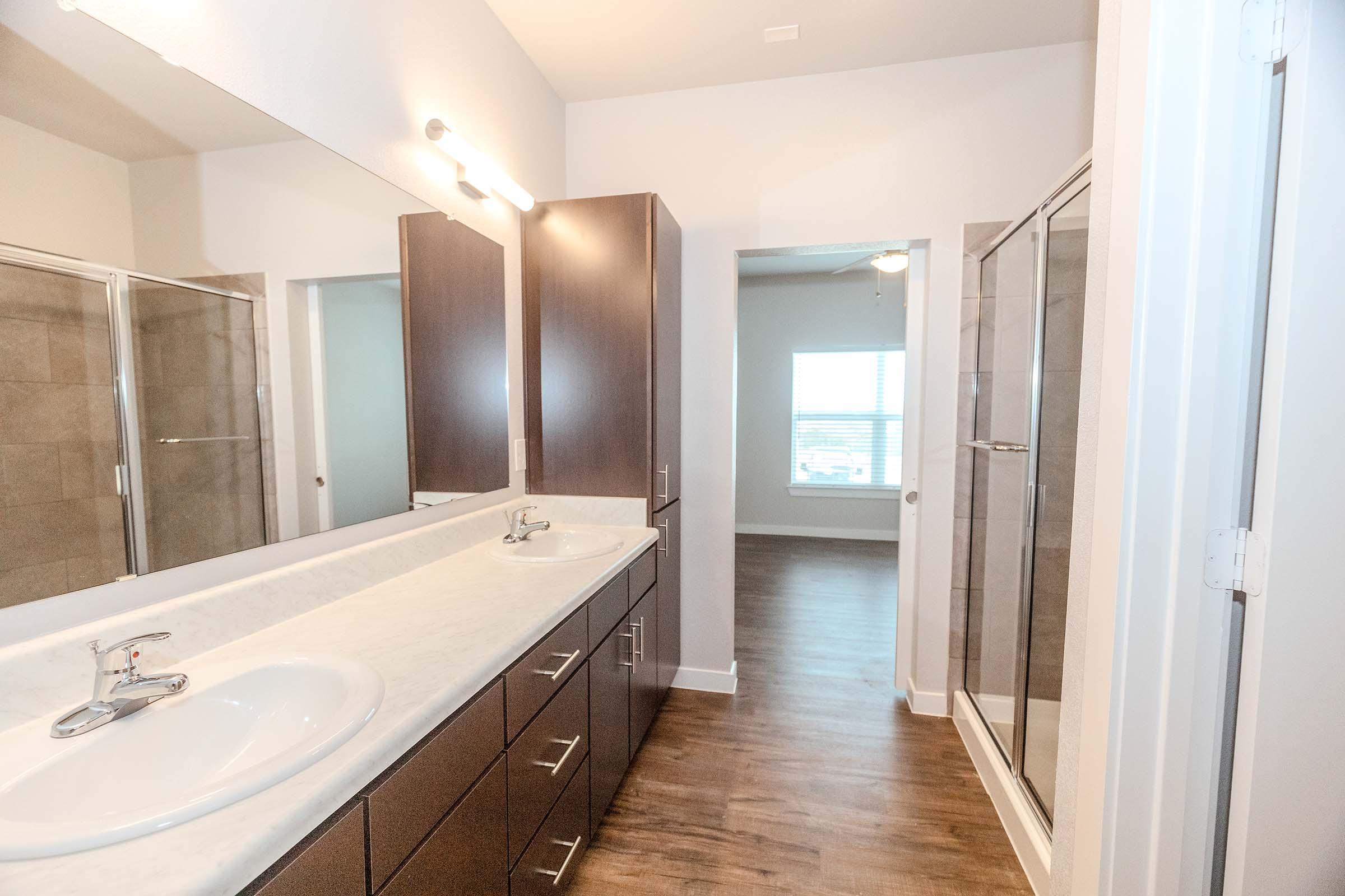 A modern bathroom featuring a dual sink vanity with a marble countertop, dark wooden cabinets, a large glass shower, and a mirror above the sinks. The space has neutral-colored walls and faux wood flooring, with a doorway leading to a well-lit room in the background. Natural light streams in through a window.