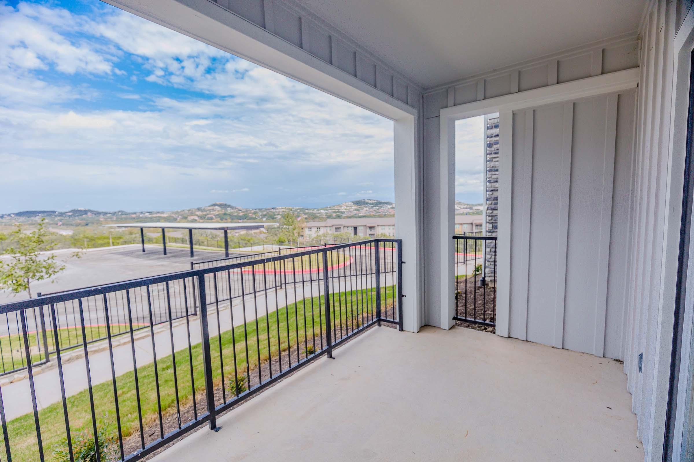 A view of a balcony with a black metal railing overlooking a landscape of hills and buildings under a partly cloudy sky. The balcony features a beige concrete floor and is attached to a modern, light-colored building.