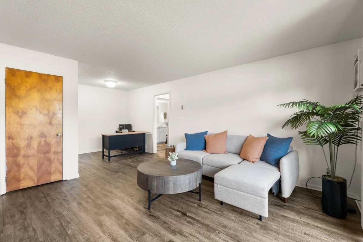 A spacious living room featuring a light gray sectional sofa with blue and orange pillows, a round coffee table, and a small potted plant. The walls are painted white, and there is a wooden door leading to another room. A desk is visible in the background, with a chair and window light.
