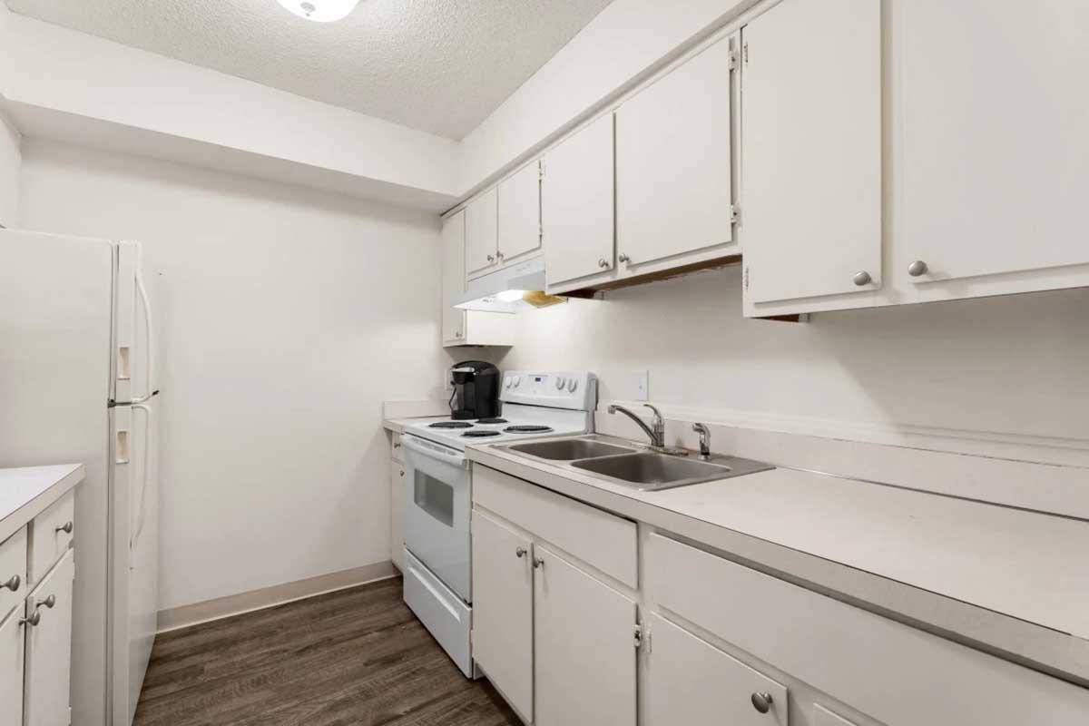 A small, modern kitchen featuring white cabinetry, a white refrigerator, and a white oven with a stovetop. There is a stainless steel sink with a countertop, and a coffee maker is placed on the countertop. The flooring is wood-like, adding a warm touch to the overall clean and functional design.