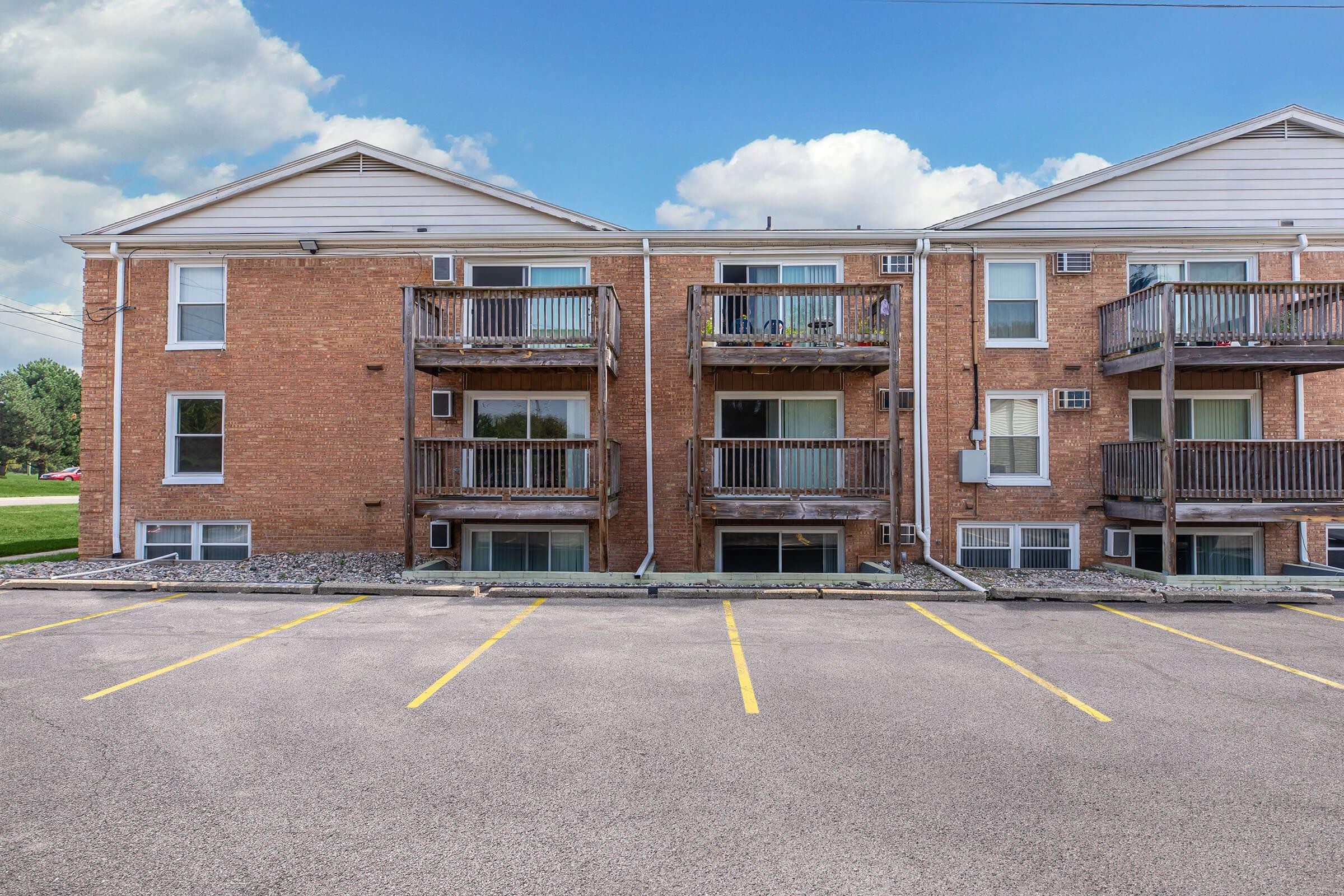A view of a brick apartment building featuring multiple balconies and windows. The building is situated in a parking lot with clearly marked yellow lines. In the background, there is a grassy area and blue sky with clouds visible.