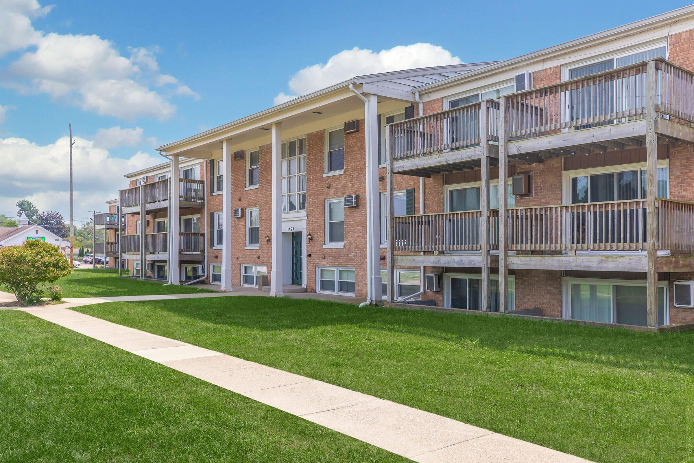 A multi-unit residential building with multiple balconies and large windows, set against a clear blue sky. The building is surrounded by well-maintained green grass and a concrete walkway leading to the entrance.