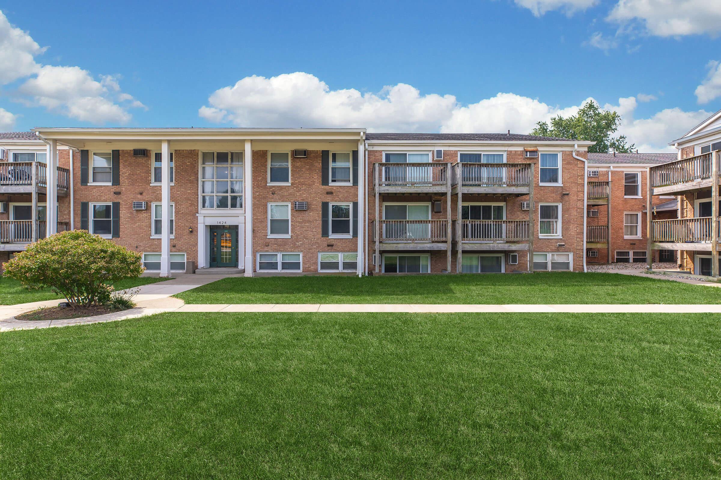 A brick apartment building with multiple balconies, surrounded by neatly trimmed grass and a sidewalk. The sky is partly cloudy, adding a bright atmosphere to the scene.