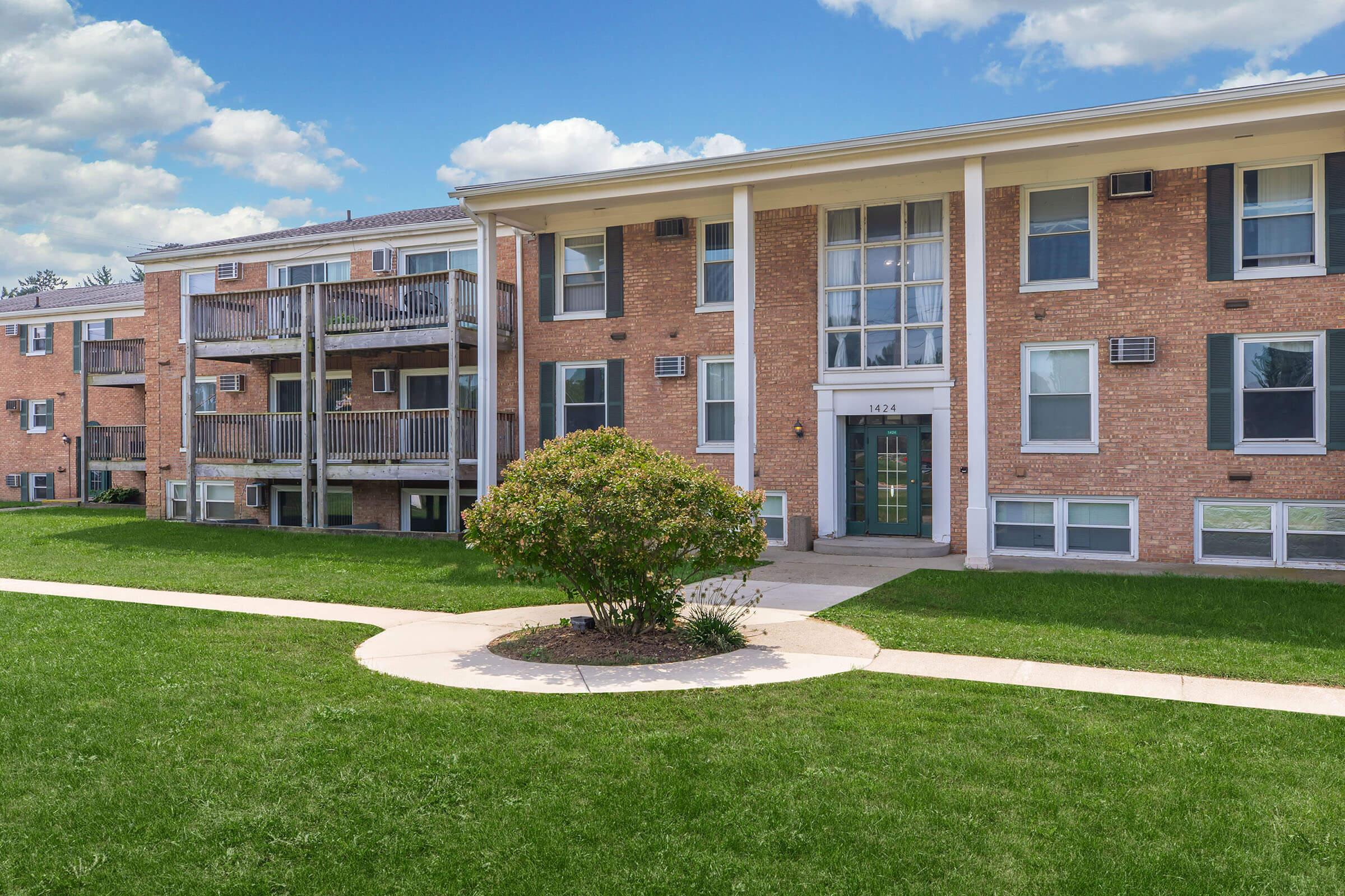 A brick apartment building with a modern entrance and large windows, surrounded by well-maintained green grass and landscaping. The building features balconies on the side, and a clear blue sky with clouds is visible above.