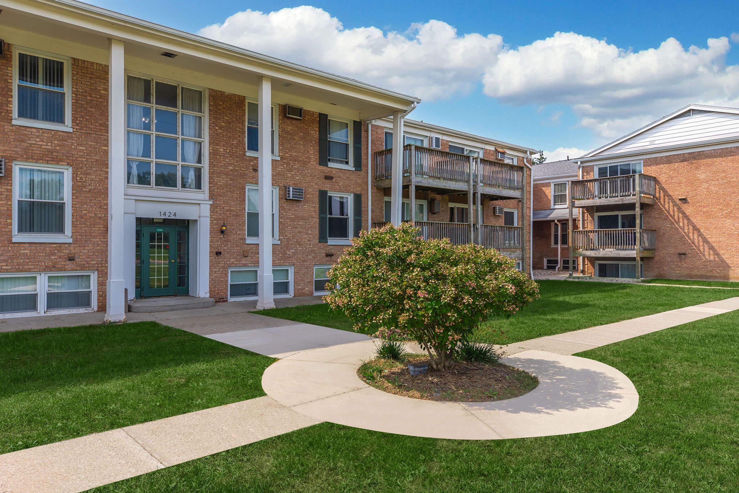 A well-maintained brick apartment complex featuring two buildings, with a landscaped garden area in the foreground. The grassy lawn surrounds a circular pathway that leads to the entrance of the main building, and there are balconies visible on the adjacent structure under a blue sky with fluffy clouds.
