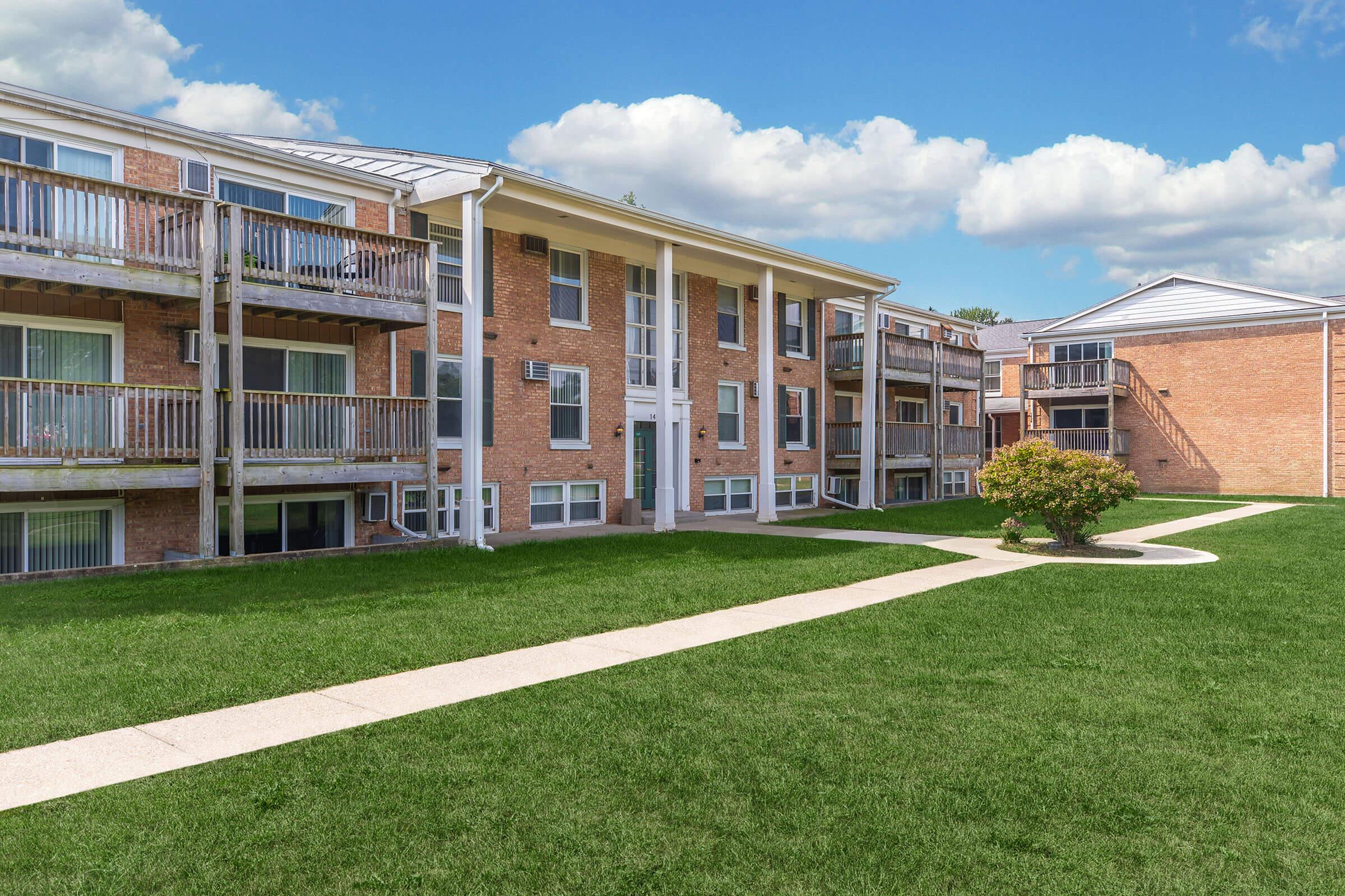 A view of a multi-story brick apartment building with balconies, surrounded by a well-maintained green lawn. A paved walkway meanders through the grass, leading to the entrance, with blue skies and fluffy clouds in the background.