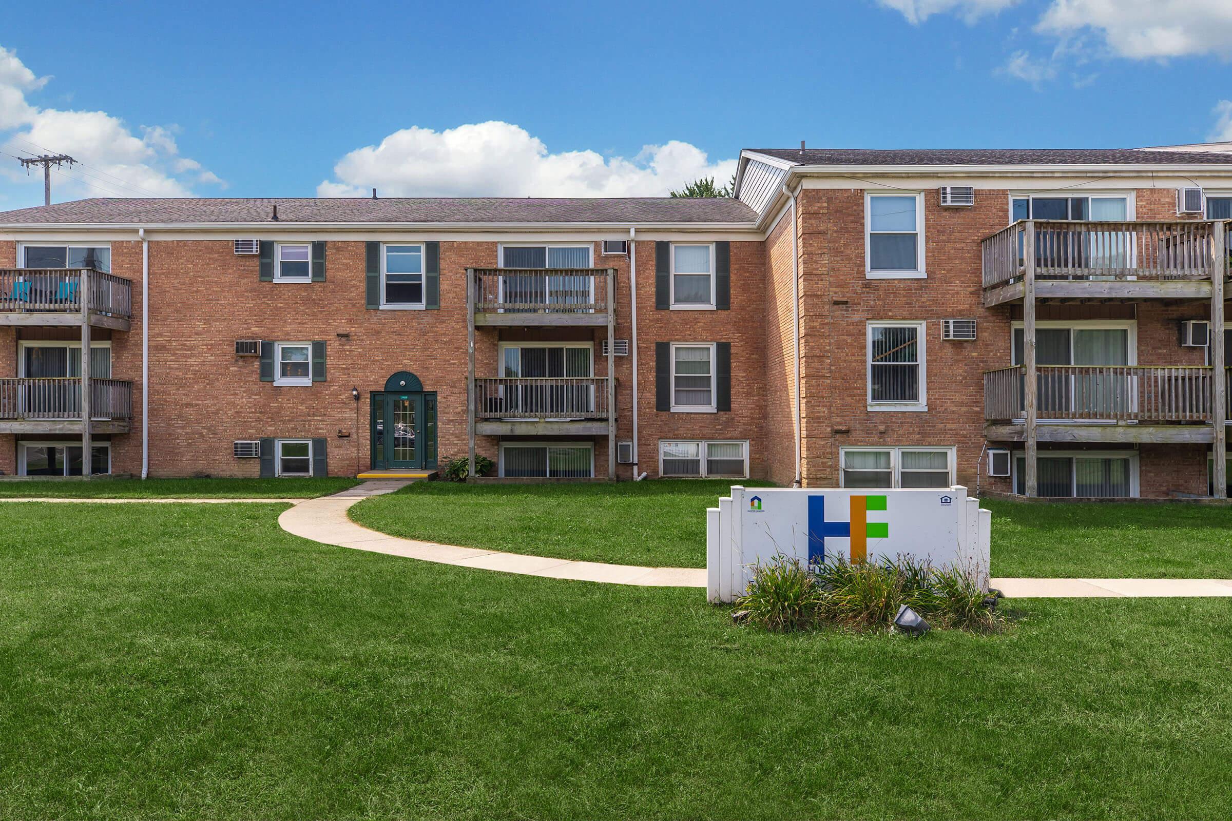 A view of a brick apartment building with several balconies, set in a green lawn area. A sign with colorful letters is visible in the foreground, indicating the name of the complex. The sky is clear with a few clouds, creating a bright, inviting atmosphere.