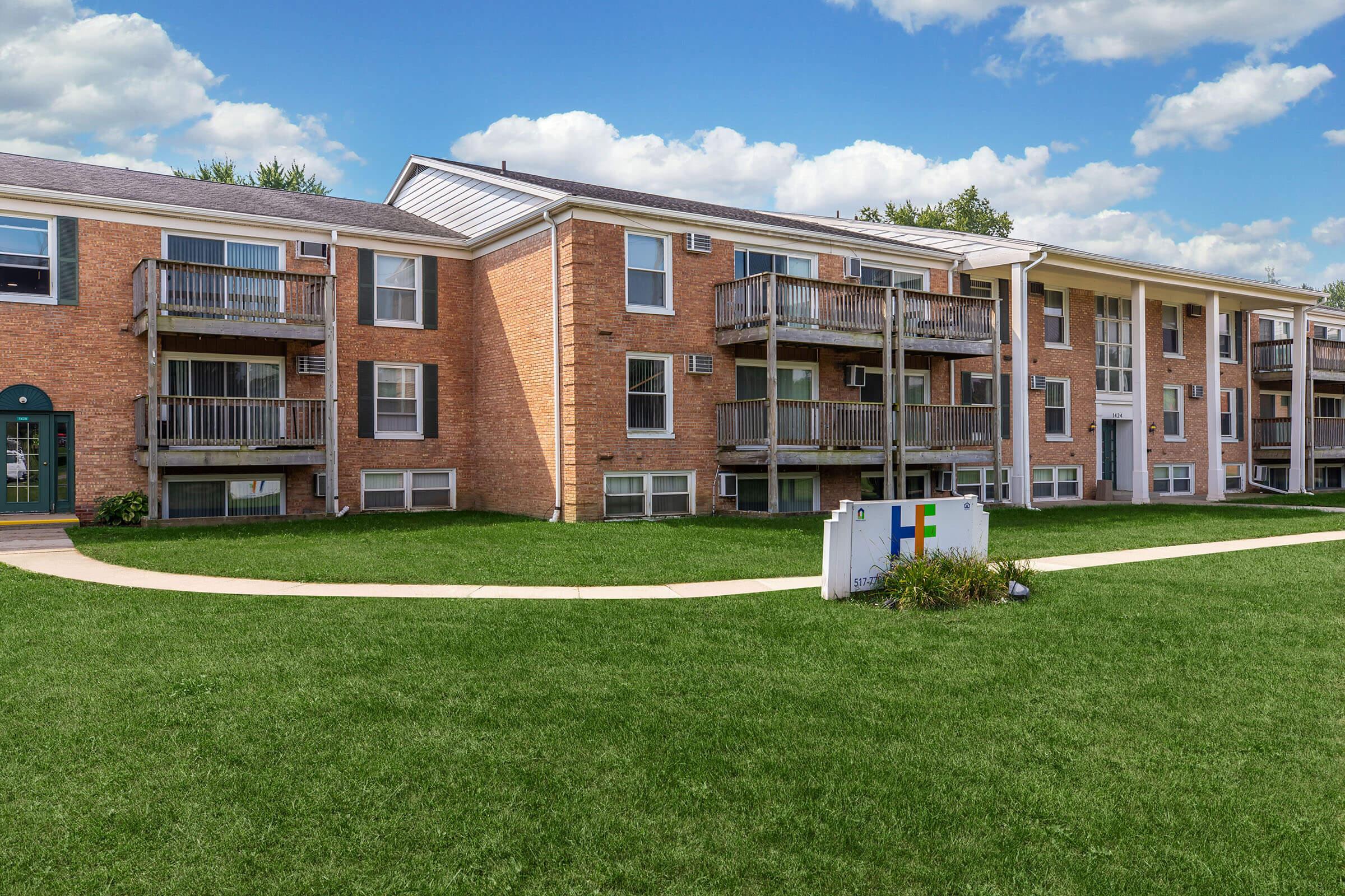A multi-unit brick apartment building with multiple balconies, set in a grassy area. The building features a central entrance and large windows. A sign indicating "Holiday Inn Express" is visible in the foreground, along with a blue sky and clouds overhead.