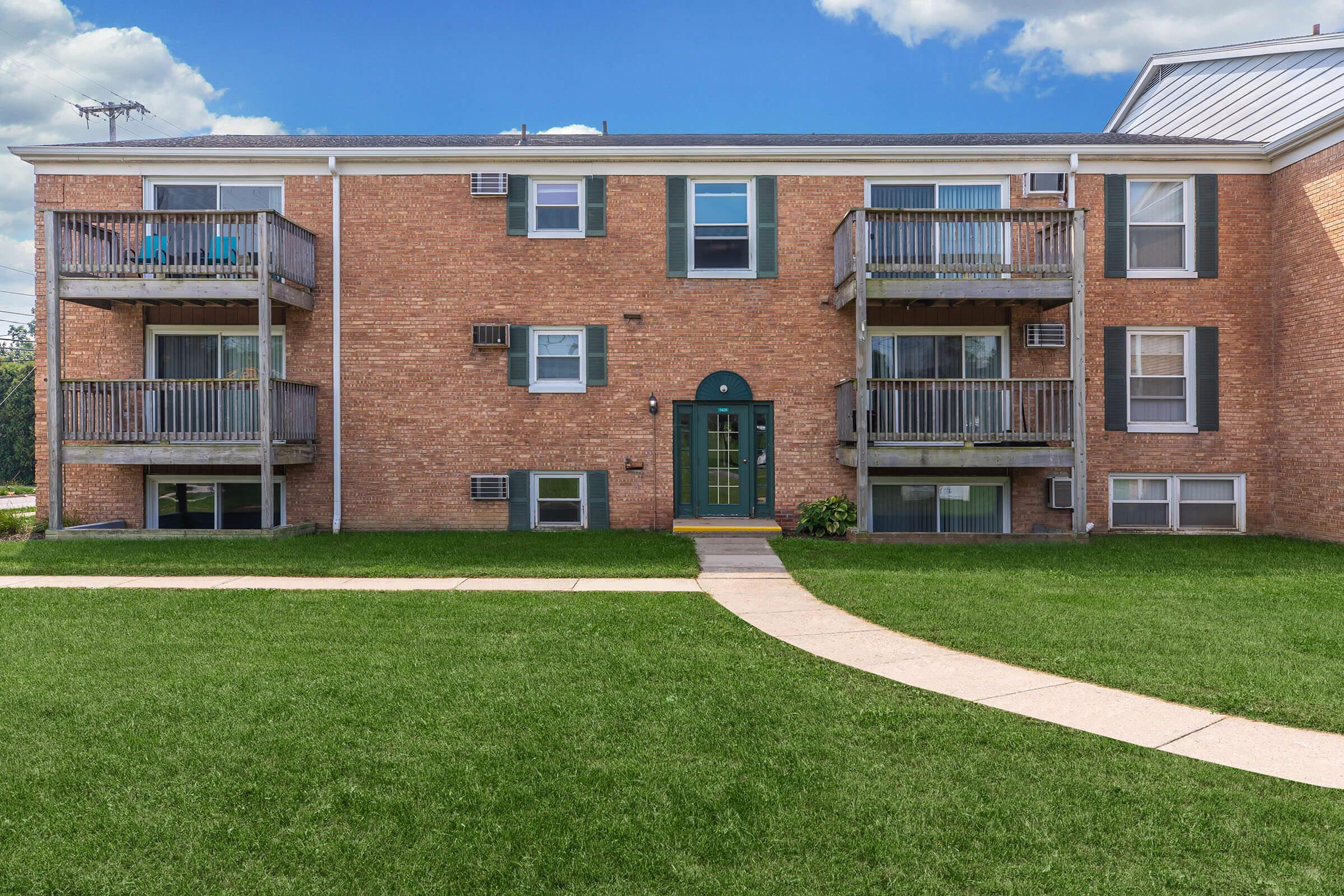 A brick apartment building featuring two levels, with balconies on the upper floor. A neat, landscaped lawn is in front, and a concrete path leads to the entrance. The sky is partly cloudy, adding to the overall inviting atmosphere of the property.