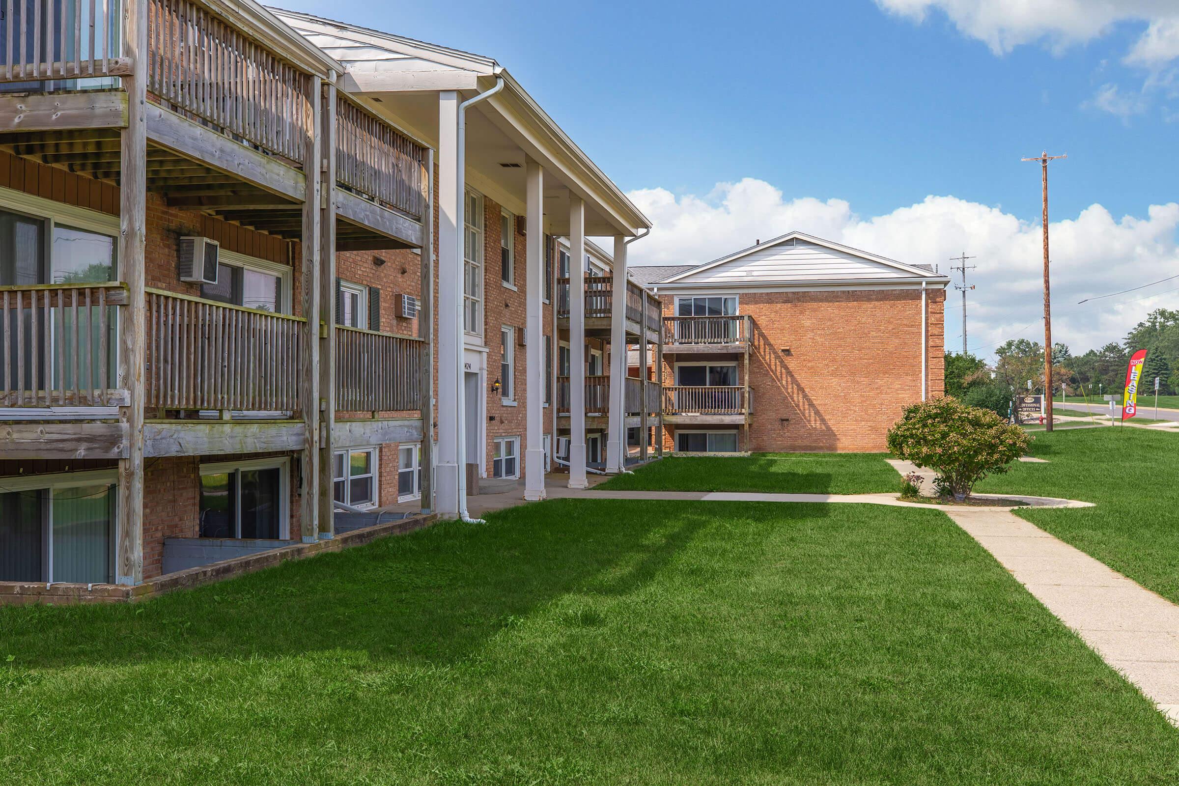 A view of two apartment buildings with balconies, surrounded by well-maintained green grass. The building on the left features multiple patios, while the right building has a modern design. The sky is clear with a few clouds, and a sidewalk leads through the grassy area.