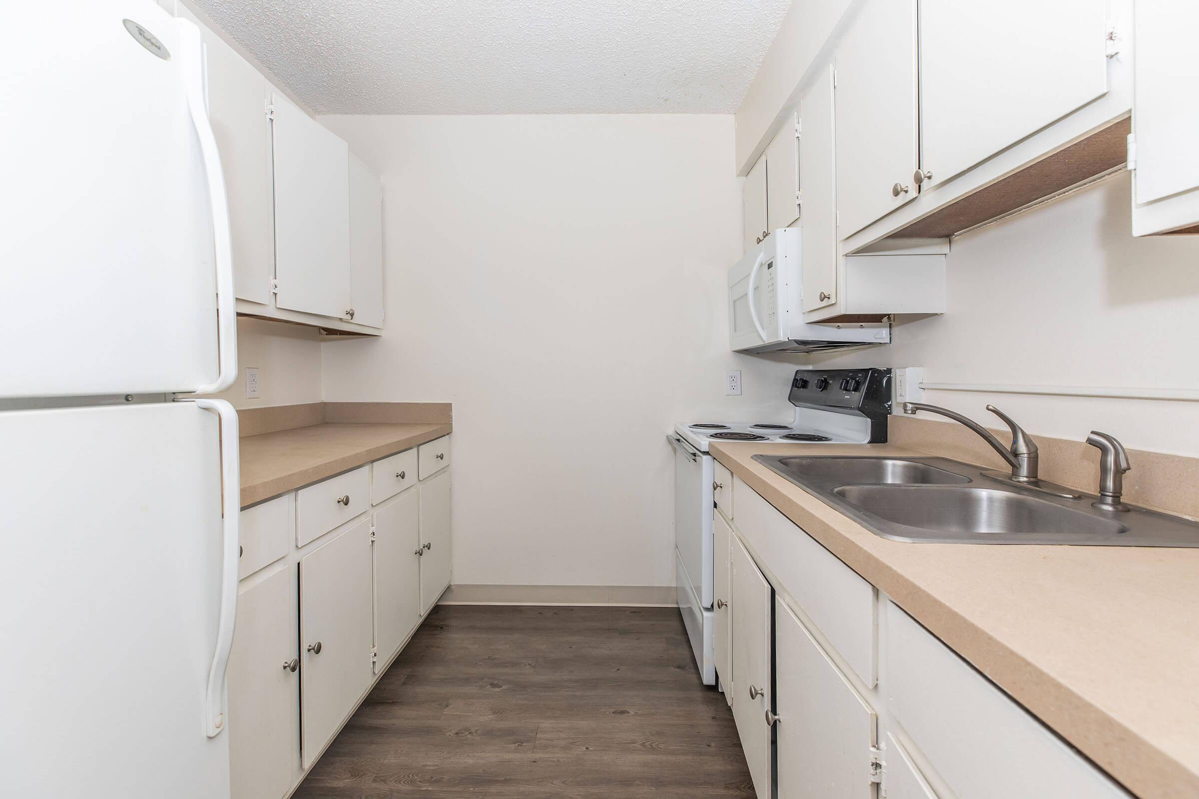 A small, modern kitchen with white cabinets and appliances. The layout includes a refrigerator, microwave, stovetop, and double sink. Light-colored countertops complement the cabinets, and the floor features a wood-like finish. The walls are painted a neutral color, creating a bright and clean atmosphere.
