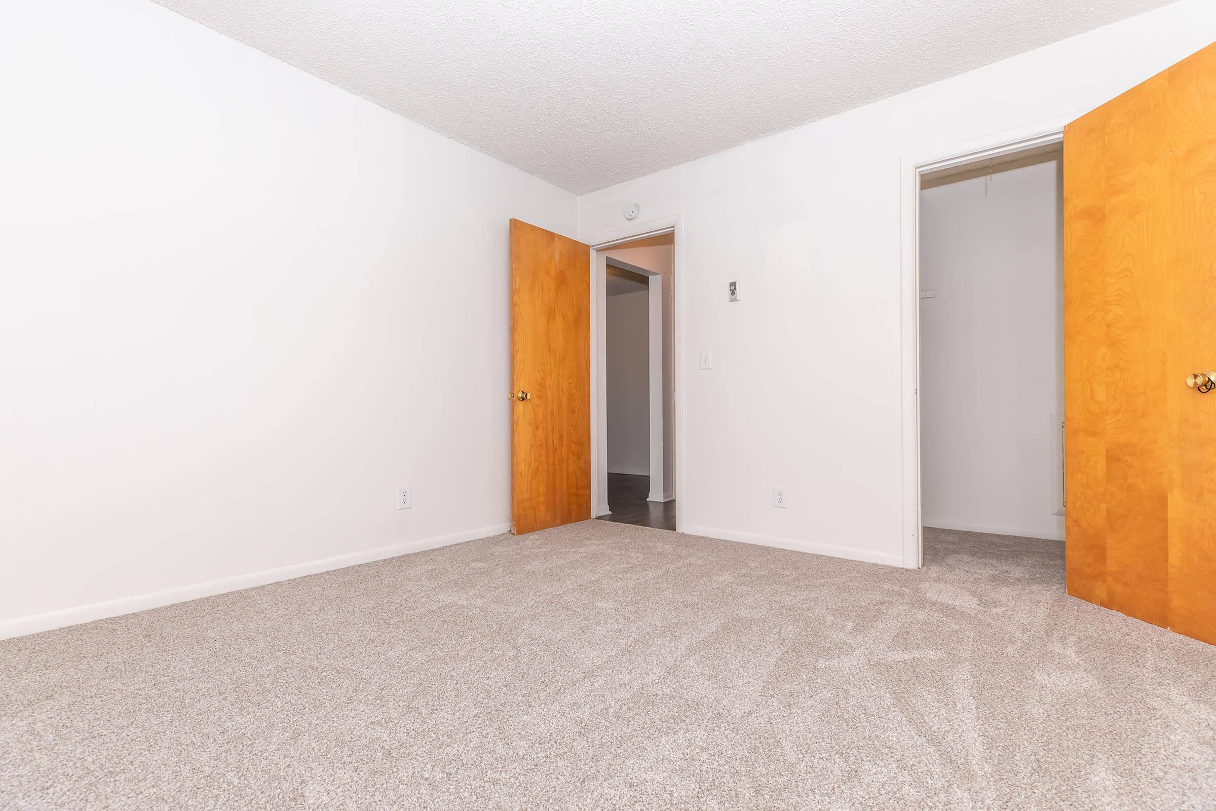 A spacious room with light-colored walls and carpet. Two wooden doors lead to adjacent areas, one slightly ajar. The room appears empty and well-lit, with no furniture, creating a clean and inviting atmosphere. The floor is carpeted and the ceiling has a textured finish.