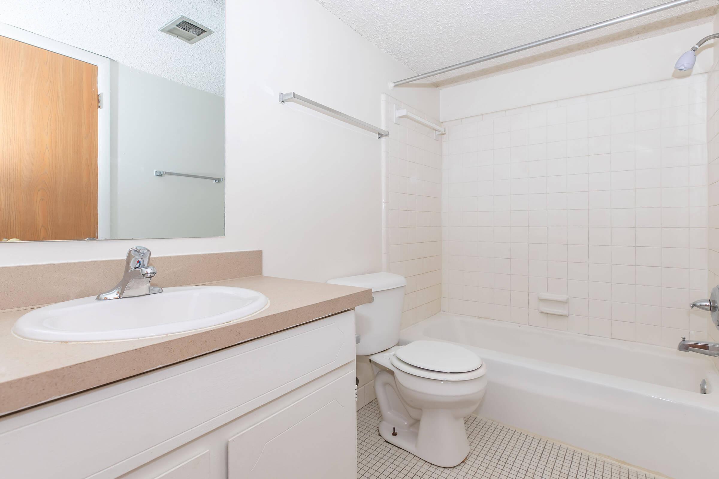 A clean, simple bathroom featuring a bathtub with a showerhead, a white toilet, a sink with a single faucet, and a large mirror above the sink. The walls are light-colored tile, and the floor has small square tiles. A wooden door is visible in the background.