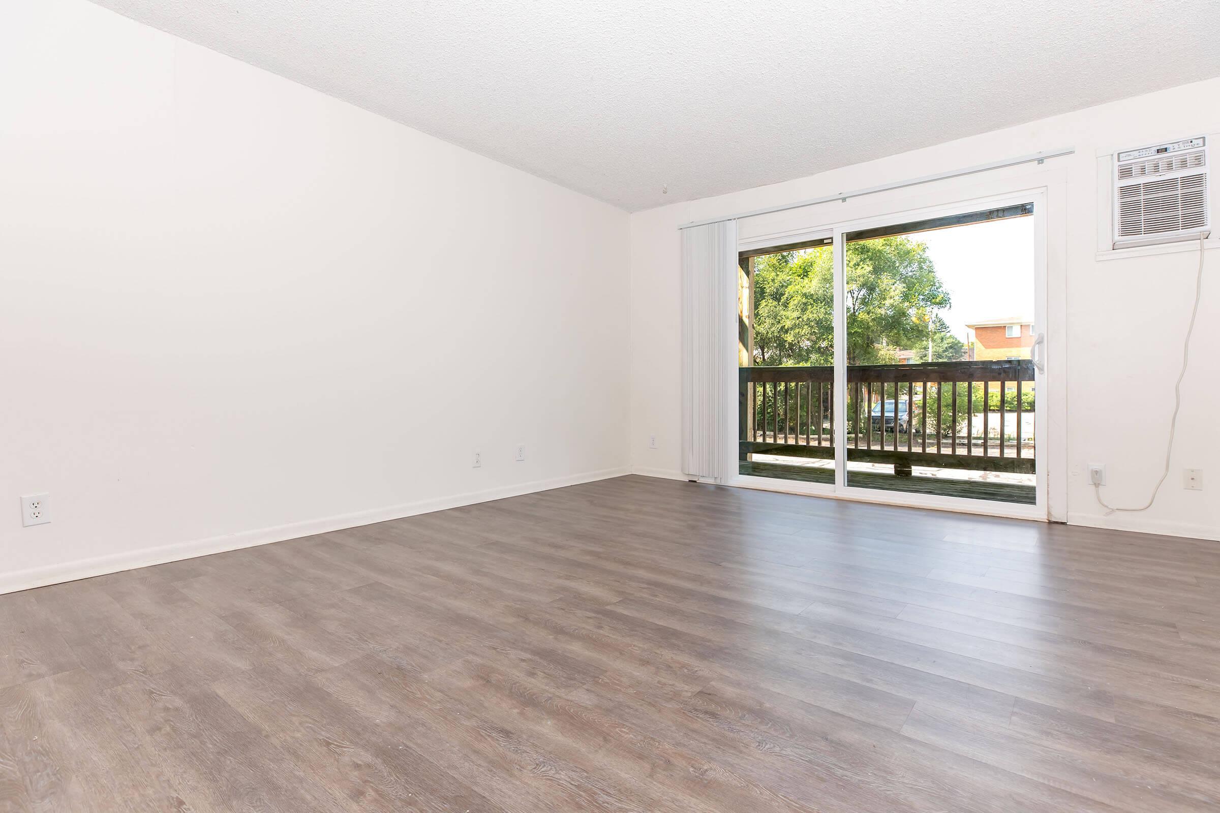 A spacious, empty room with light-colored walls and a large window that opens to a balcony. The floor is covered in light wood laminate, and there's an air conditioning unit mounted on the wall. The overall atmosphere is bright and open, ideal for personal customization.