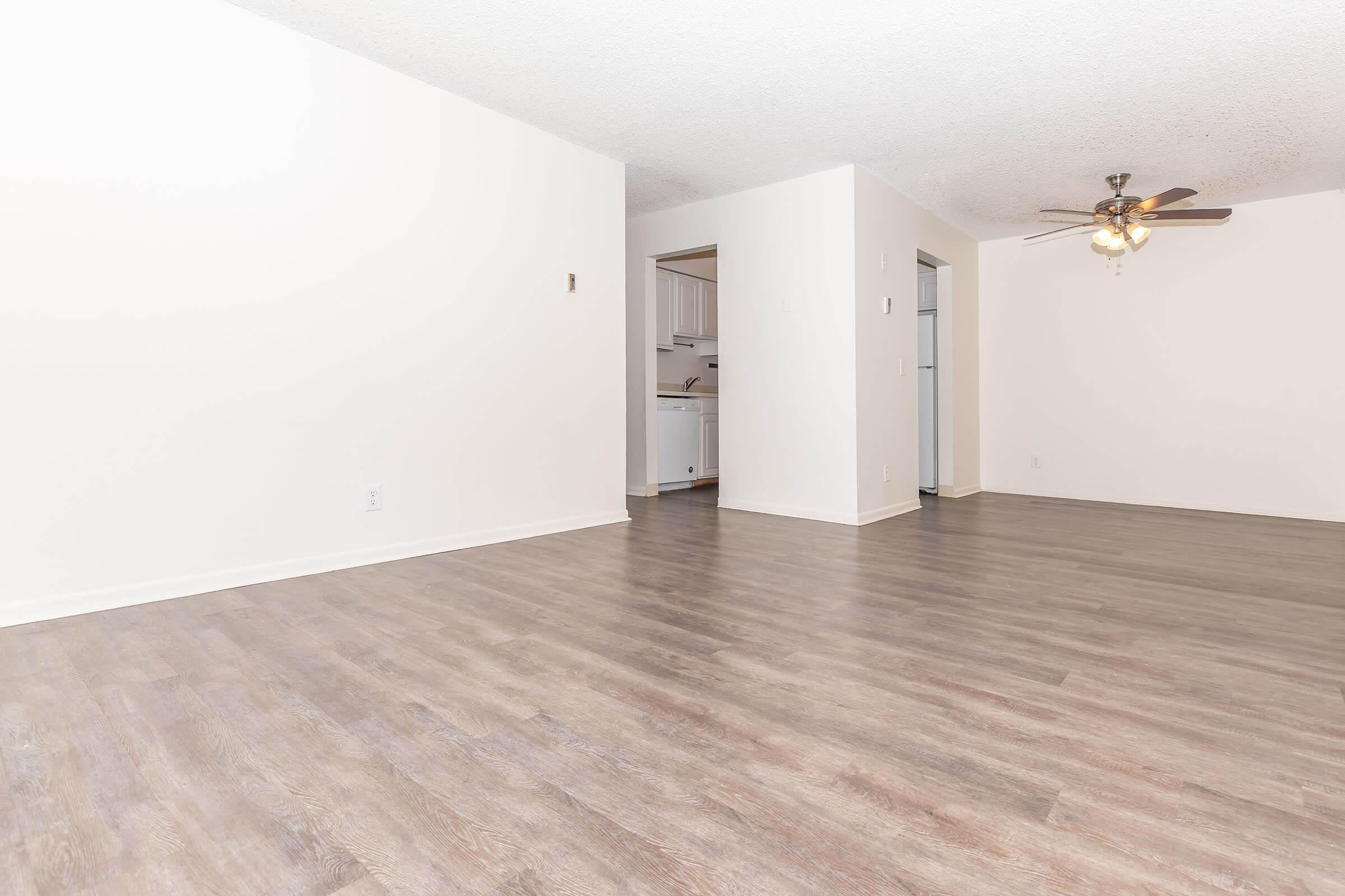 An empty room featuring light-colored walls and a wood-like laminate floor. A ceiling fan is visible in the corner, and a doorway leads to another space, likely a kitchen or utility area. The overall atmosphere is bright and spacious, with natural lighting present.