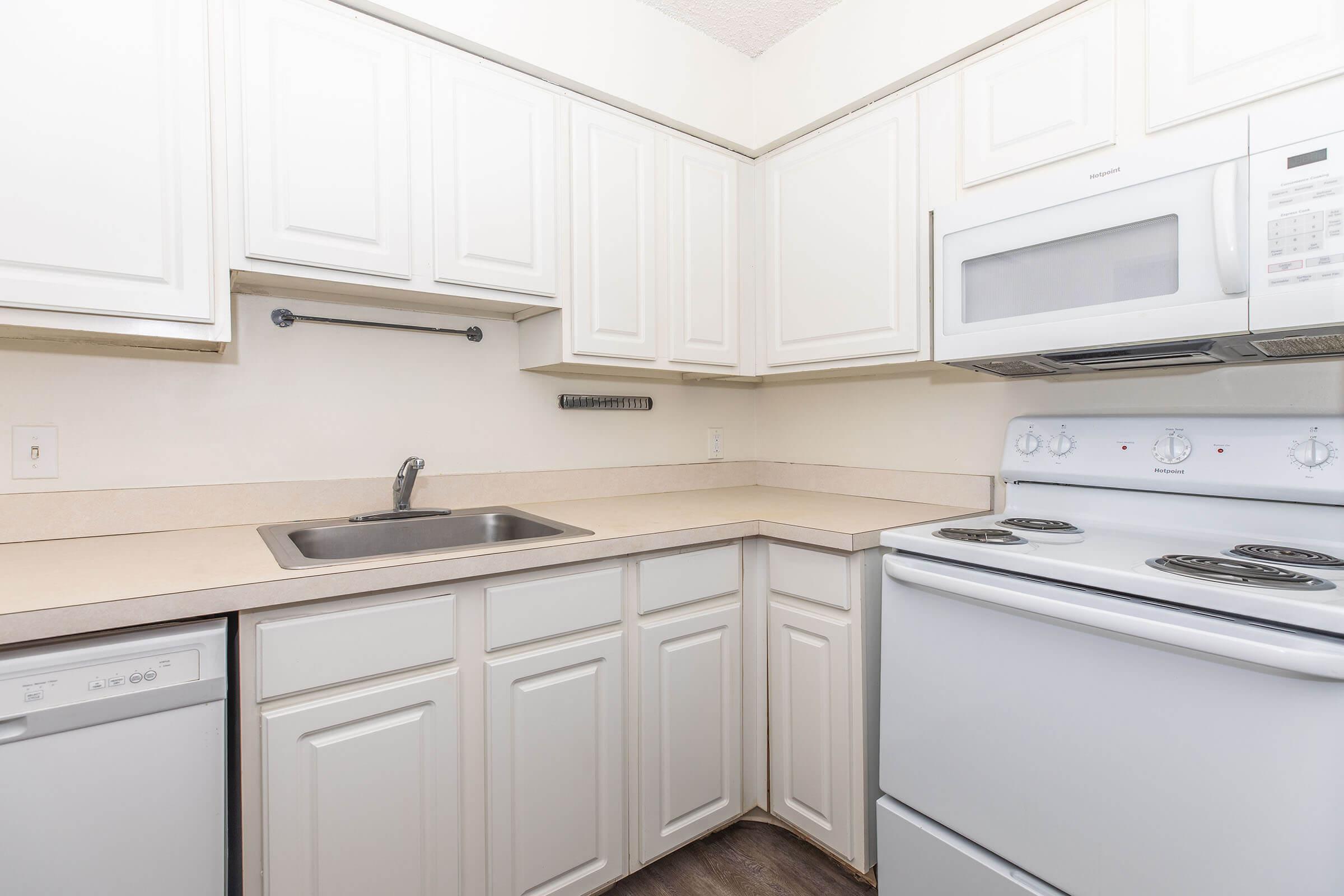 A modern kitchen featuring white cabinetry, a stainless steel sink, a dishwasher, and an electric stove with an oven. The countertop is light-colored, and there is a microwave above the stove. The design is clean and contemporary, with a neutral color scheme and minimal decor.