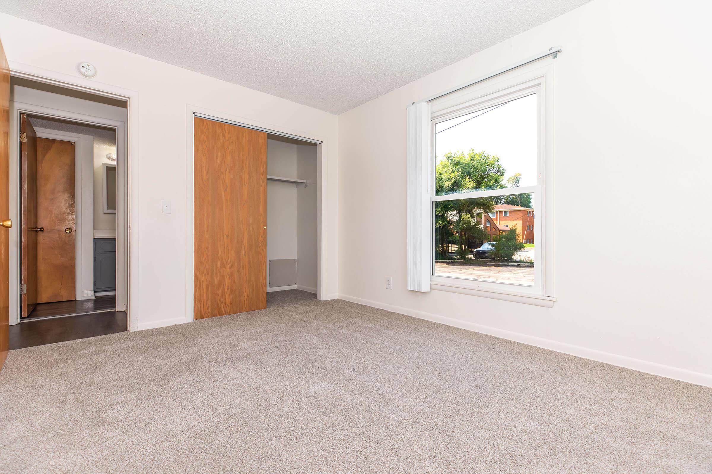 Empty room with light-colored carpet and white walls. A window allows natural light to enter, revealing a view of greenery outside. Two doors lead to closets and another room. The overall space is tidy and well-lit, creating a welcoming atmosphere.