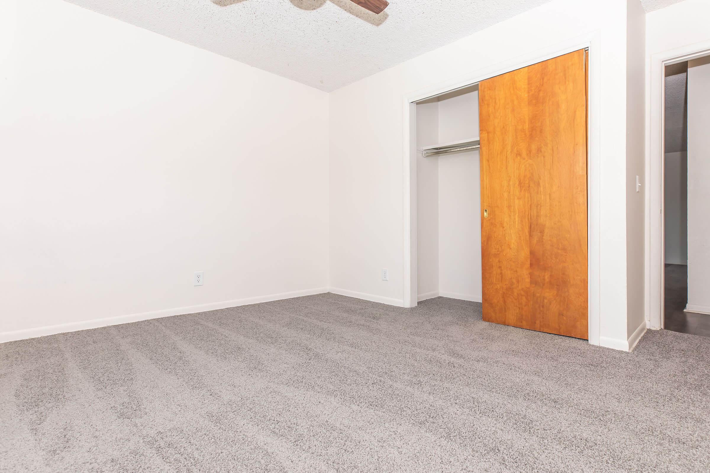 A minimal interior room featuring light-colored walls and carpet. There is a wooden closet door on the right and an open closet space, with no furniture present, creating a spacious and airy feel. A ceiling fan is visible at the top, adding to the room's features.