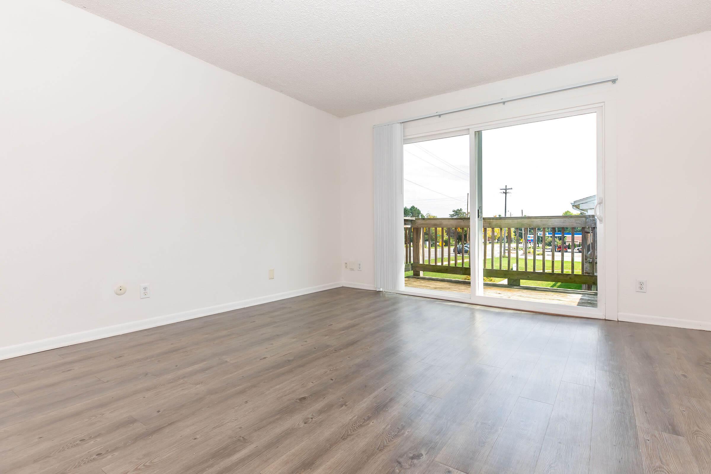 Empty room with light-colored walls and a wooden floor. A large sliding glass door leads to a balcony, allowing natural light to enter. The view outside includes a grassy area and some trees, with utility poles visible in the background. The space has a modern and minimalistic feel.