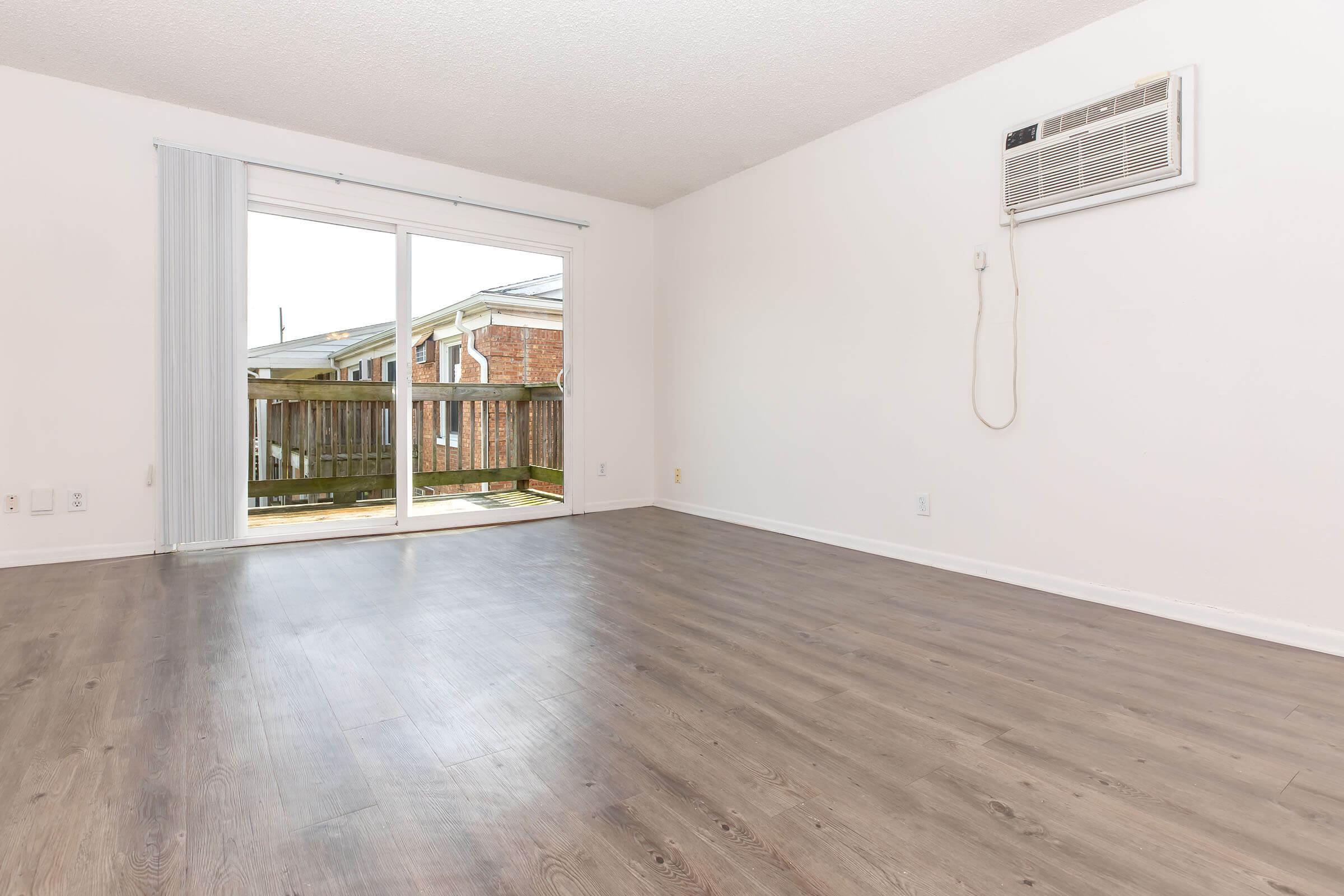 A spacious, empty room with light-colored walls and a smooth wooden floor. A sliding glass door leads to a balcony, allowing natural light into the space. An air conditioning unit is mounted above the door. The room has minimal furnishings, creating an open and airy feel.