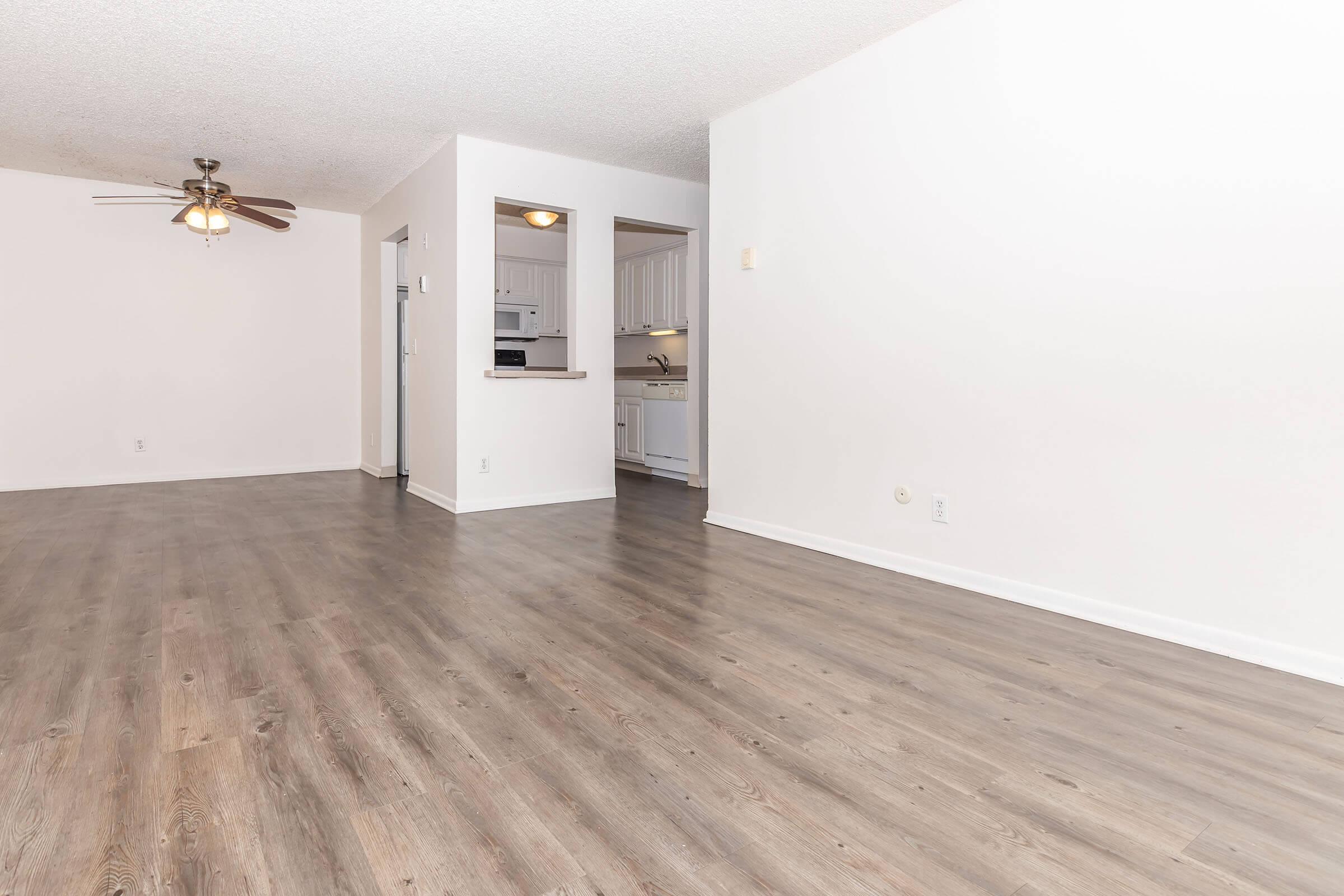 Spacious living area with light-colored walls and a ceiling fan. To the left, an open doorway leads to a kitchen with white cabinetry and appliances. The flooring is a modern laminate in a natural wood finish, enhancing the bright, airy feel of the space.