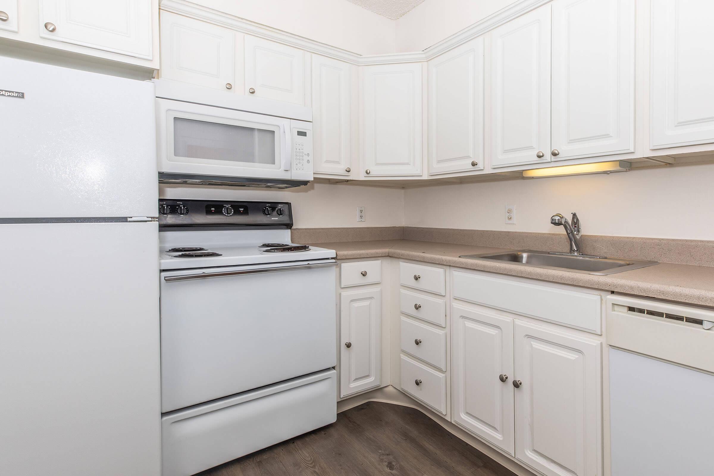 A modern kitchen featuring white cabinetry, a white refrigerator, a built-in microwave above a stove with four burners, and a single sink. The countertops are beige, complementing the cabinetry, and there is a light fixture above the sink for illumination. The flooring is dark wood.