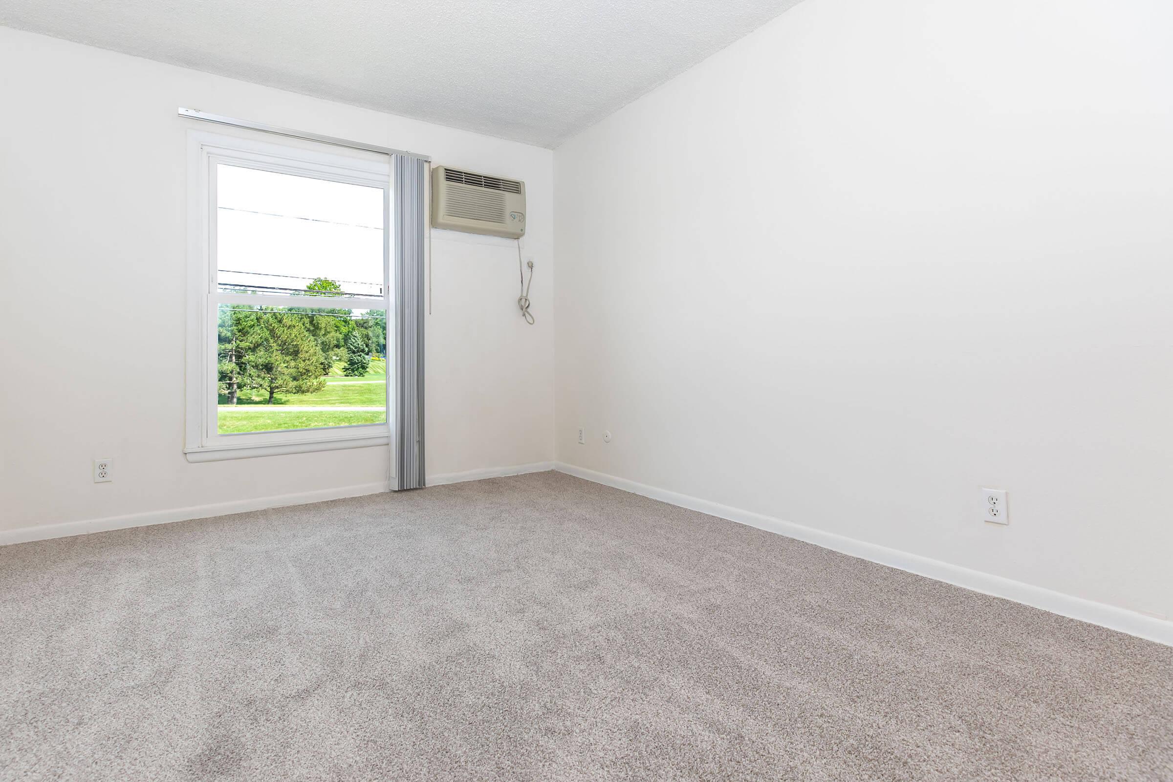 A spacious, empty room with neutral-colored walls and plush carpet. There's a window with light gray curtains, showing a view of greenery outside. An air conditioning unit is mounted on the wall, and the sunlight brightens the space, creating a clean and inviting atmosphere.