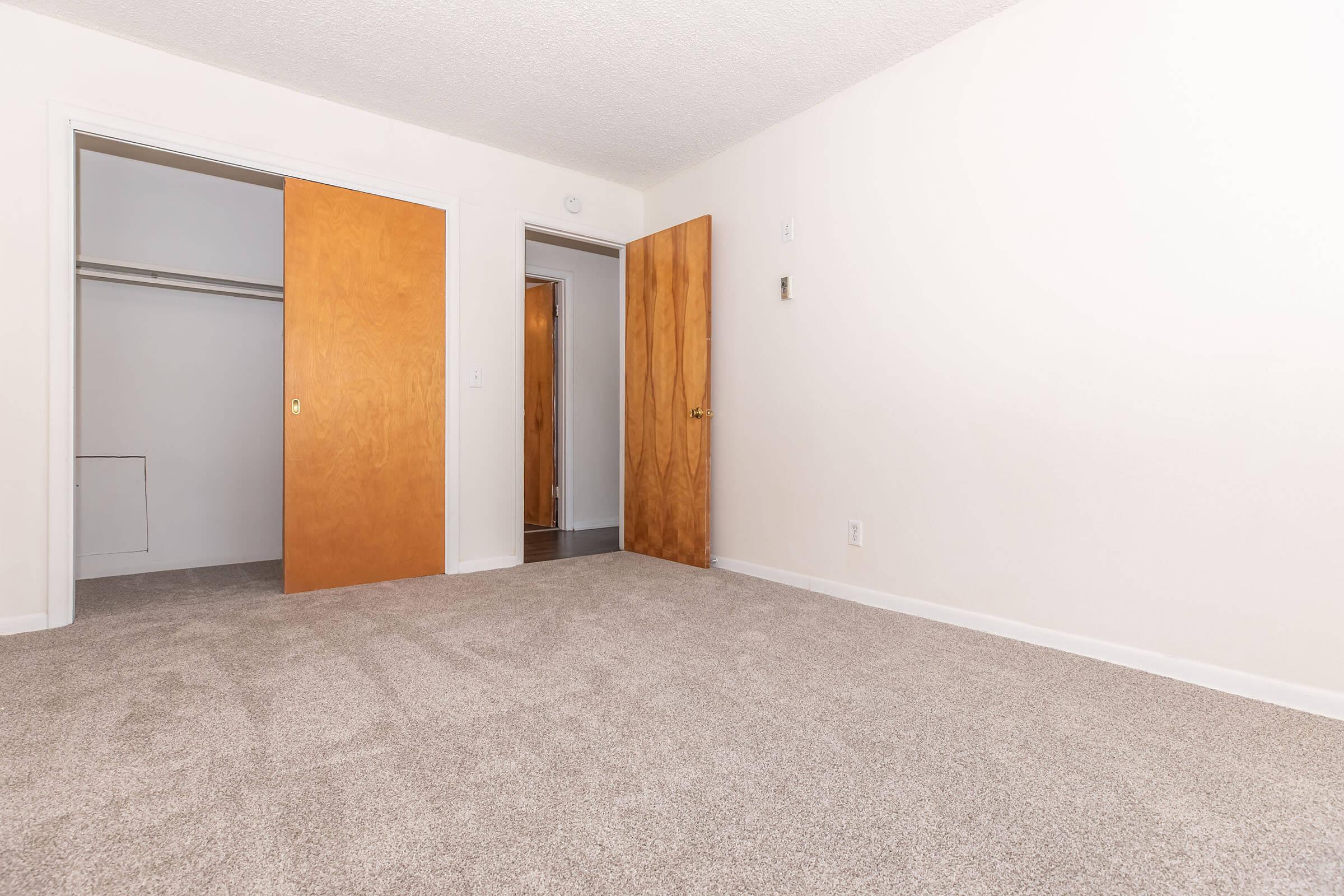 A spacious and empty room featuring light-colored walls and new carpeting. Two wooden doors lead to closets and another room, providing access and storage options. Natural light fills the area, highlighting the clean and simple design, making it suitable for various uses.