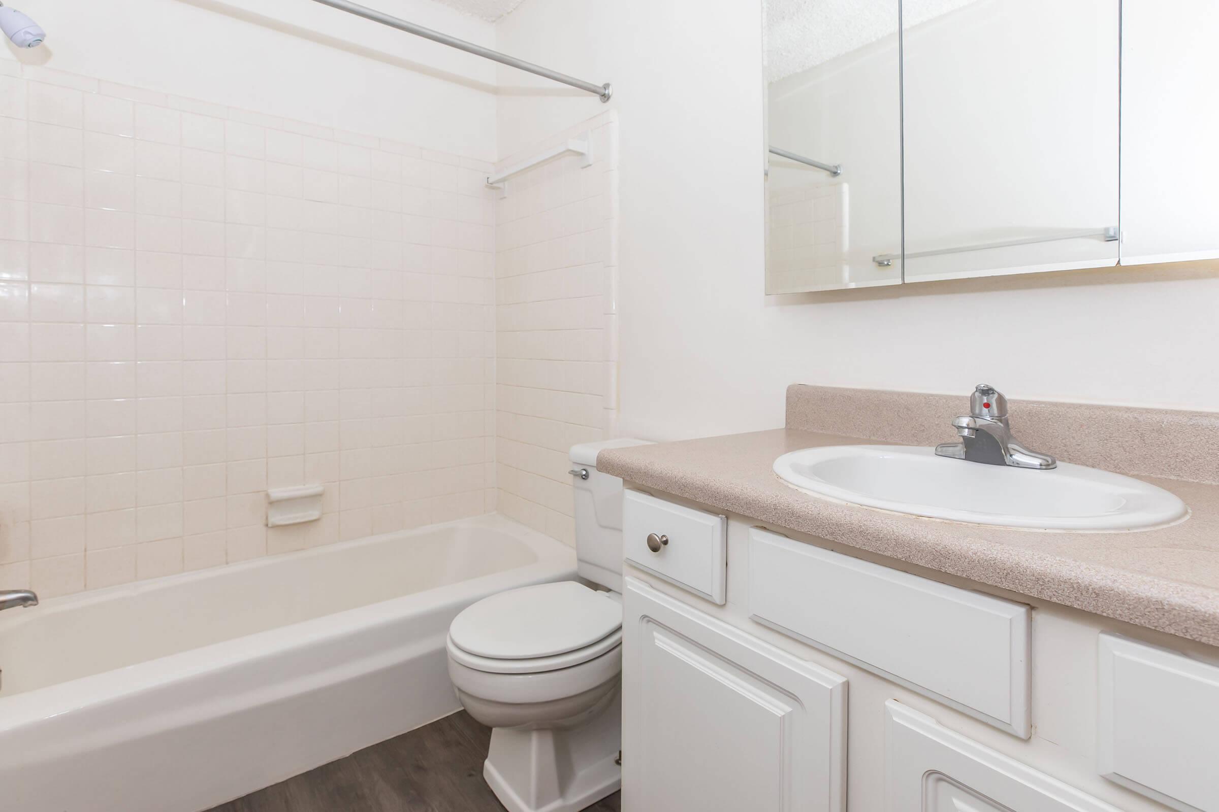 A clean and simple bathroom featuring a bathtub with a showerhead, a toilet, and a sink with a mirror above. The walls are white with light-colored tiles, and the flooring is a dark, wood-like finish. The aesthetic is bright and minimalist, emphasizing space and cleanliness.