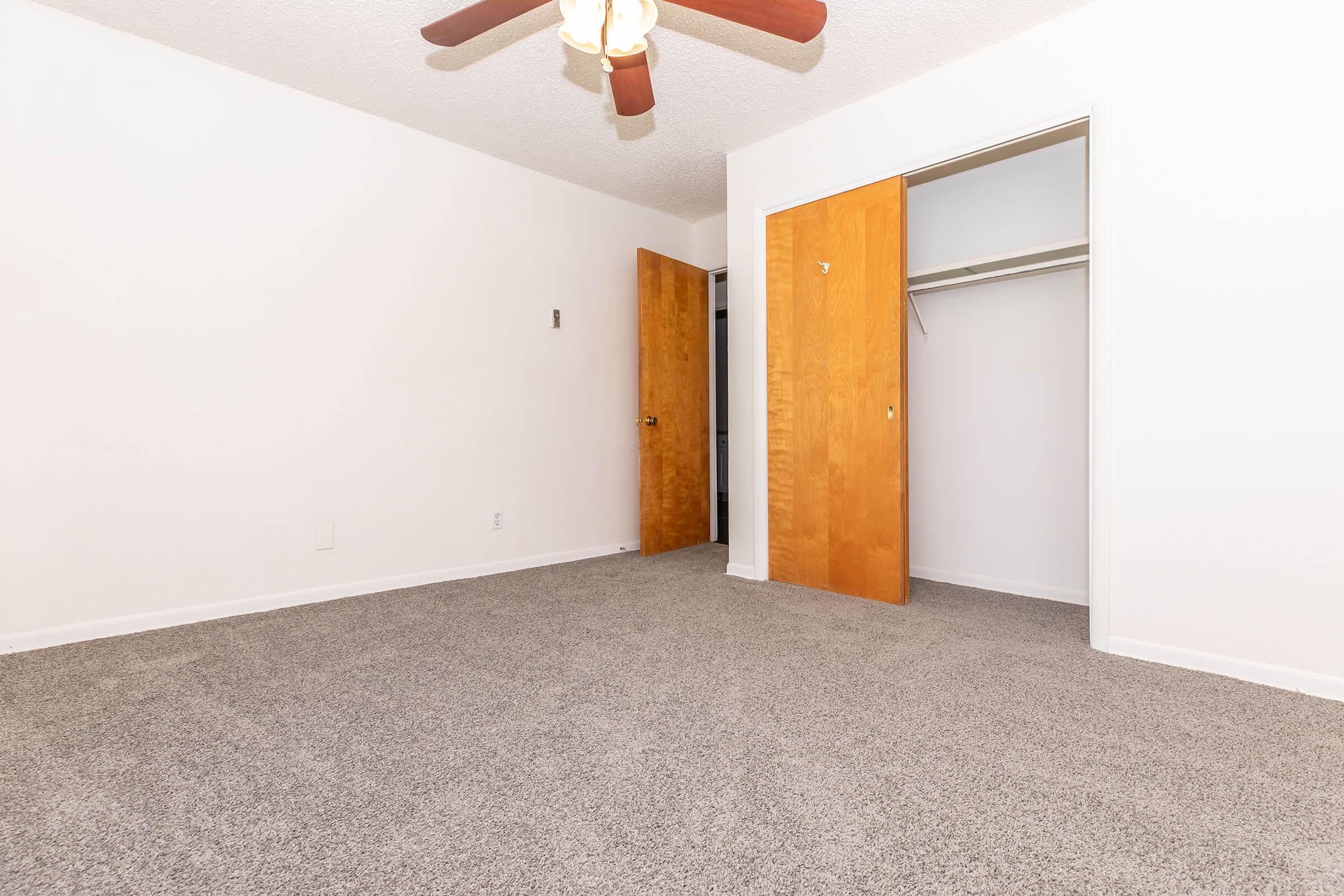 A view of a vacant room with light-colored walls and carpet. There is a ceiling fan with wooden blades, and two wooden doors are slightly ajar, leading to a closet and another space. The room is well-lit and has no furniture, emphasizing the open space.